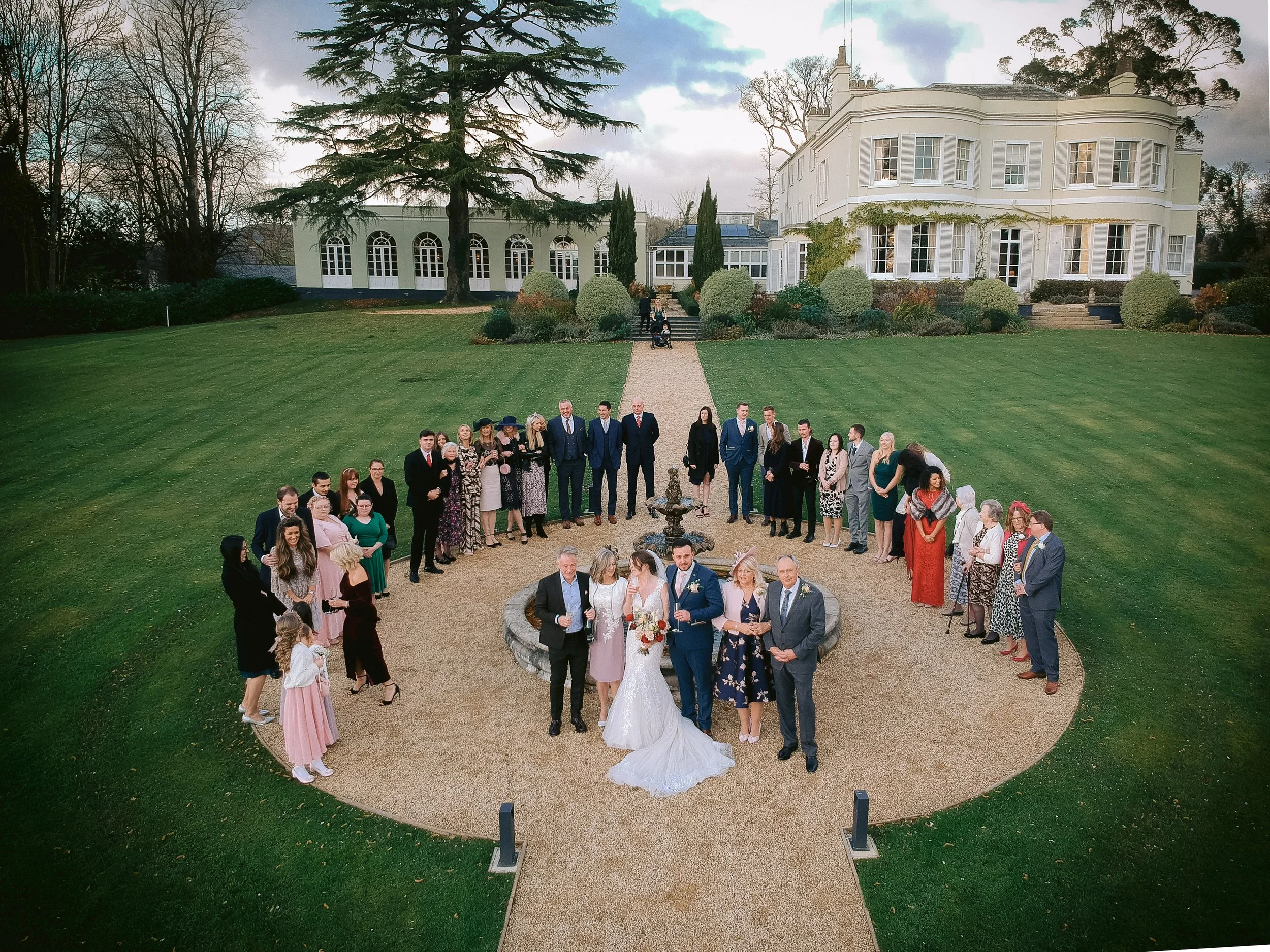 Group of wedding guests gathered around bride and groom outdoors in front of a large white house with a manicured lawn.