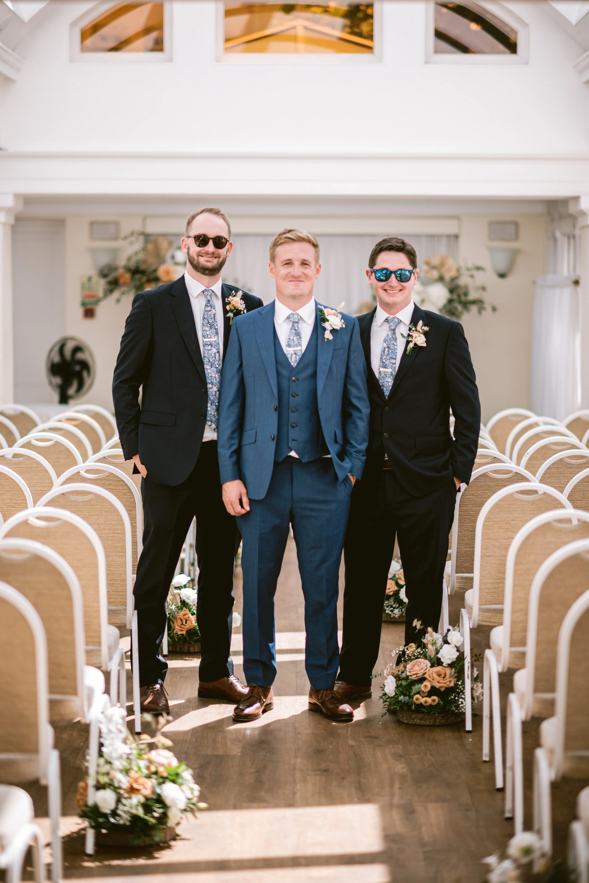 Three men in suits standing inside a decorated wedding venue with chairs and floral arrangements, smiling at the camera.