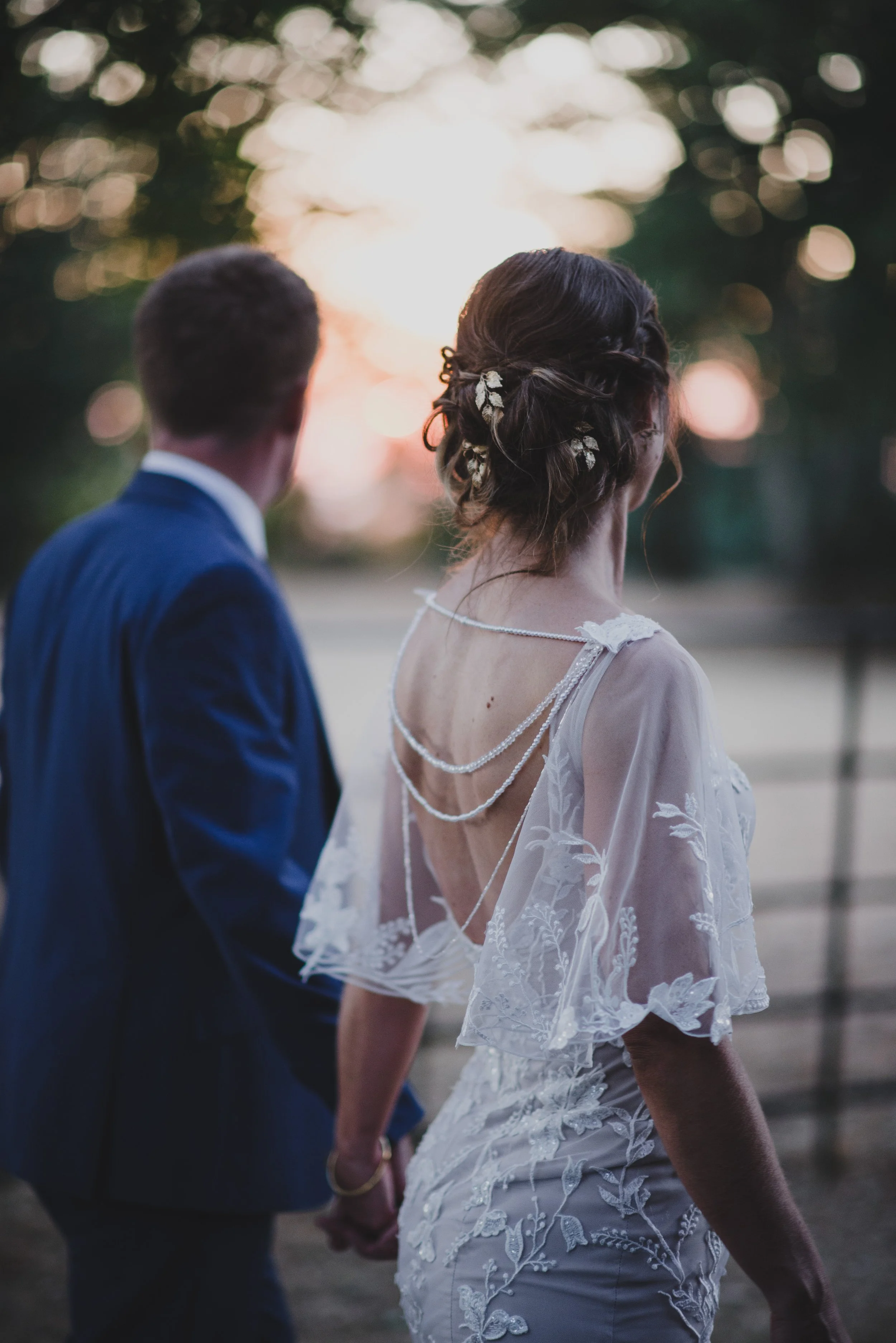 Bride and groom holding hands outdoors at sunset, with the bride wearing a lace wedding dress and decorated hair, and the groom in a blue suit.
