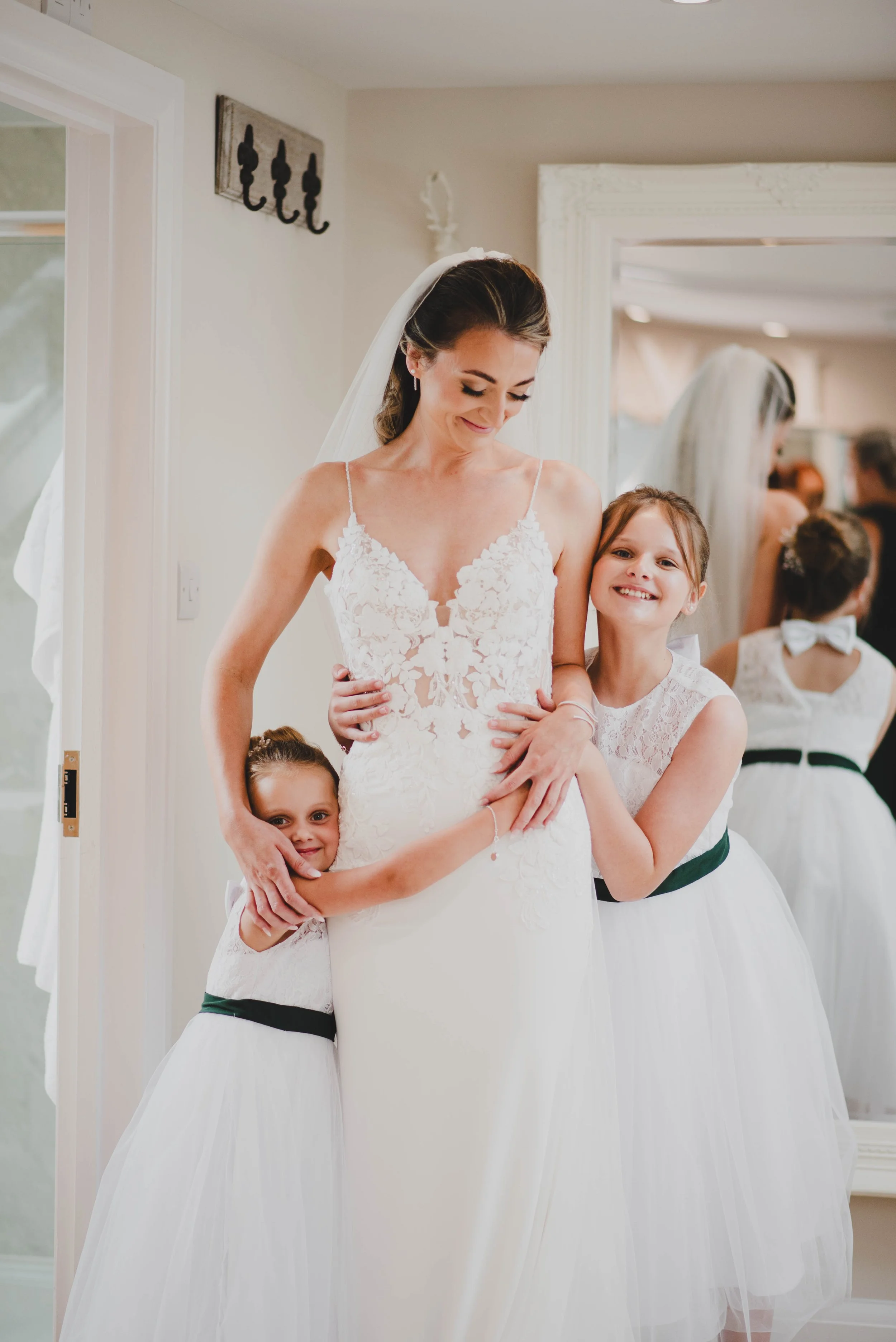 A bride in a white lace wedding dress smiling as she hugs two young girls in white dresses with black sashes, inside a room with a mirror and wall decor.
