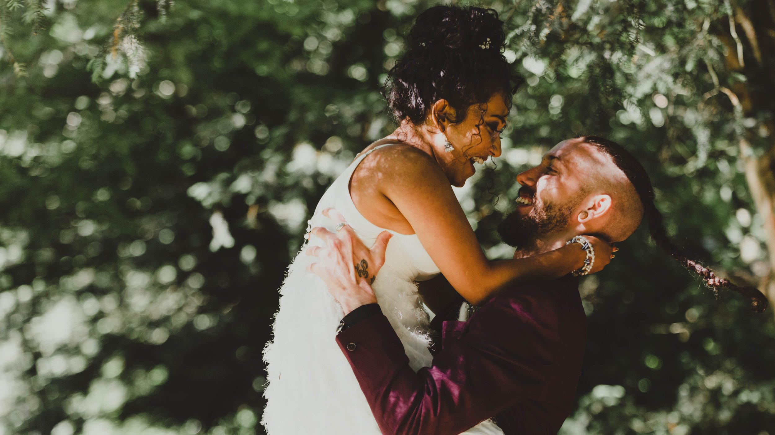 A joyful couple, a woman and a man, sharing a tender, happy moment outdoors among greenery. The woman is dressed in a white dress, and the man is wearing a dark suit. They are smiling at each other, with the woman being lifted slightly in the air.