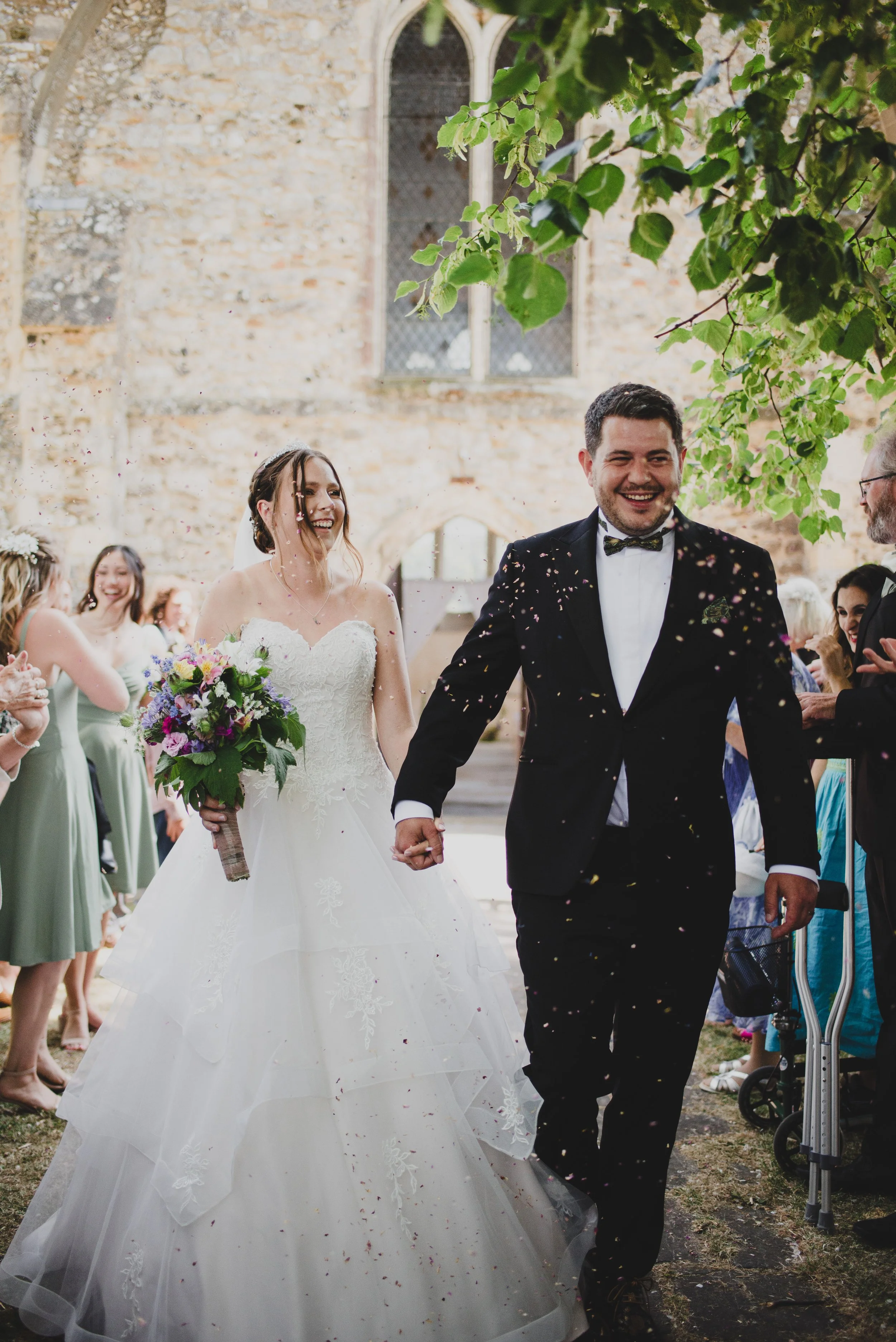 A newlywed couple walking hand in hand outside a church, smiling as confetti falls around them, with guests on either side celebrating their wedding.
