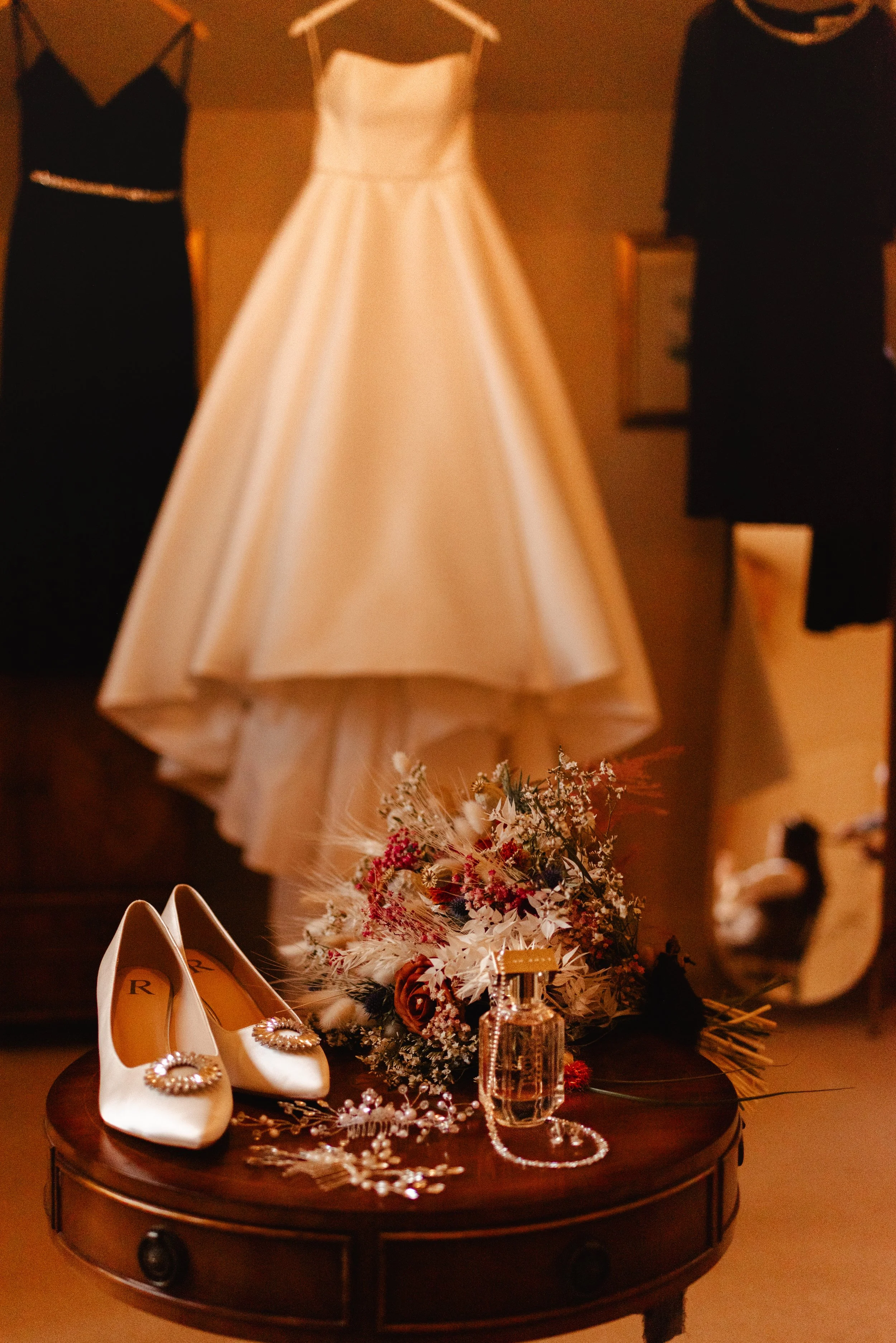 A pair of white wedding shoes with jeweled embellishments on the toes, a bouquet of flowers, perfume, and jewelry on a round wooden table. In the background, a white wedding dress and two black dresses are hanging.