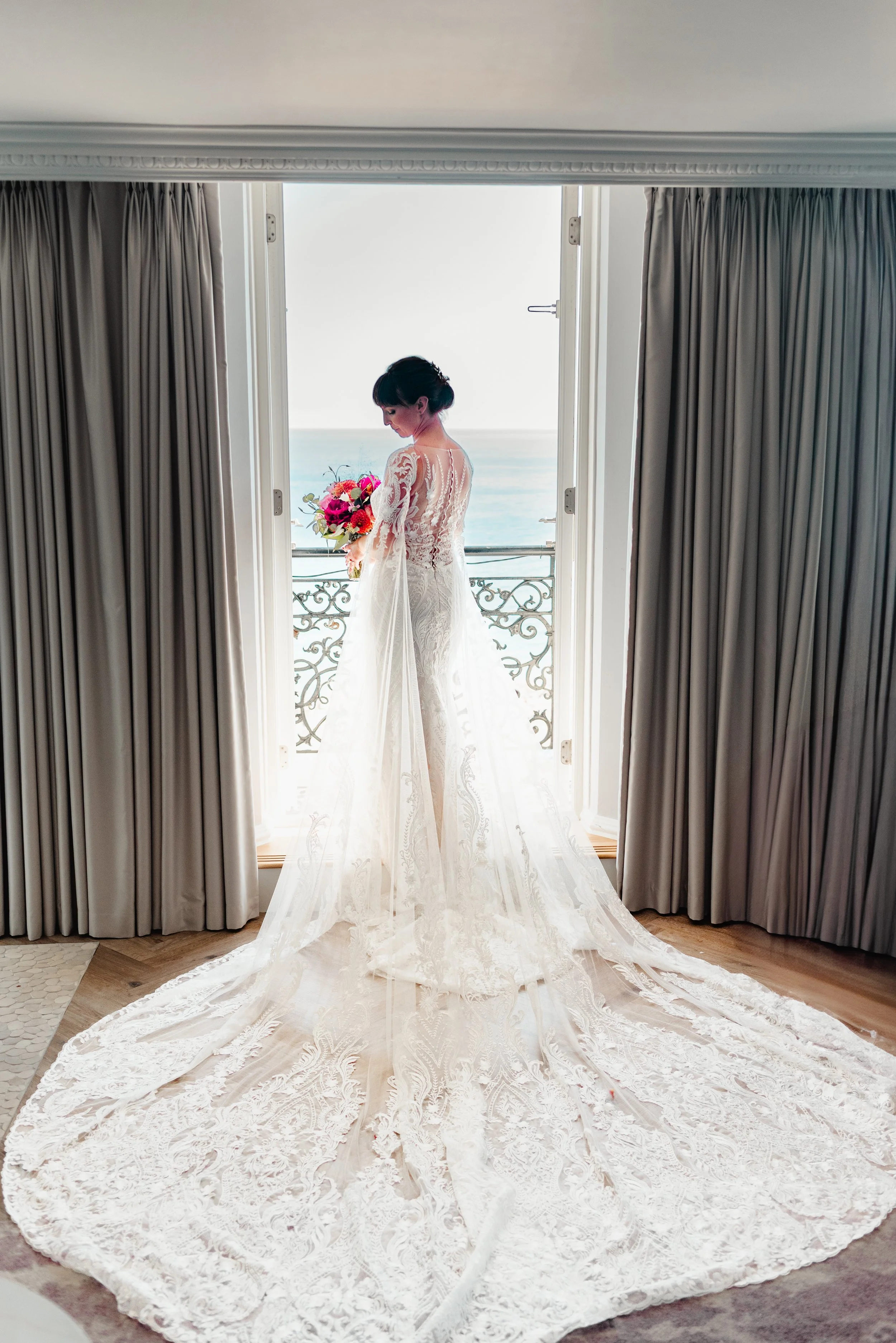 Bridal woman standing on a balcony in an elegant lace wedding dress, holding a bouquet, overlooking the ocean.
