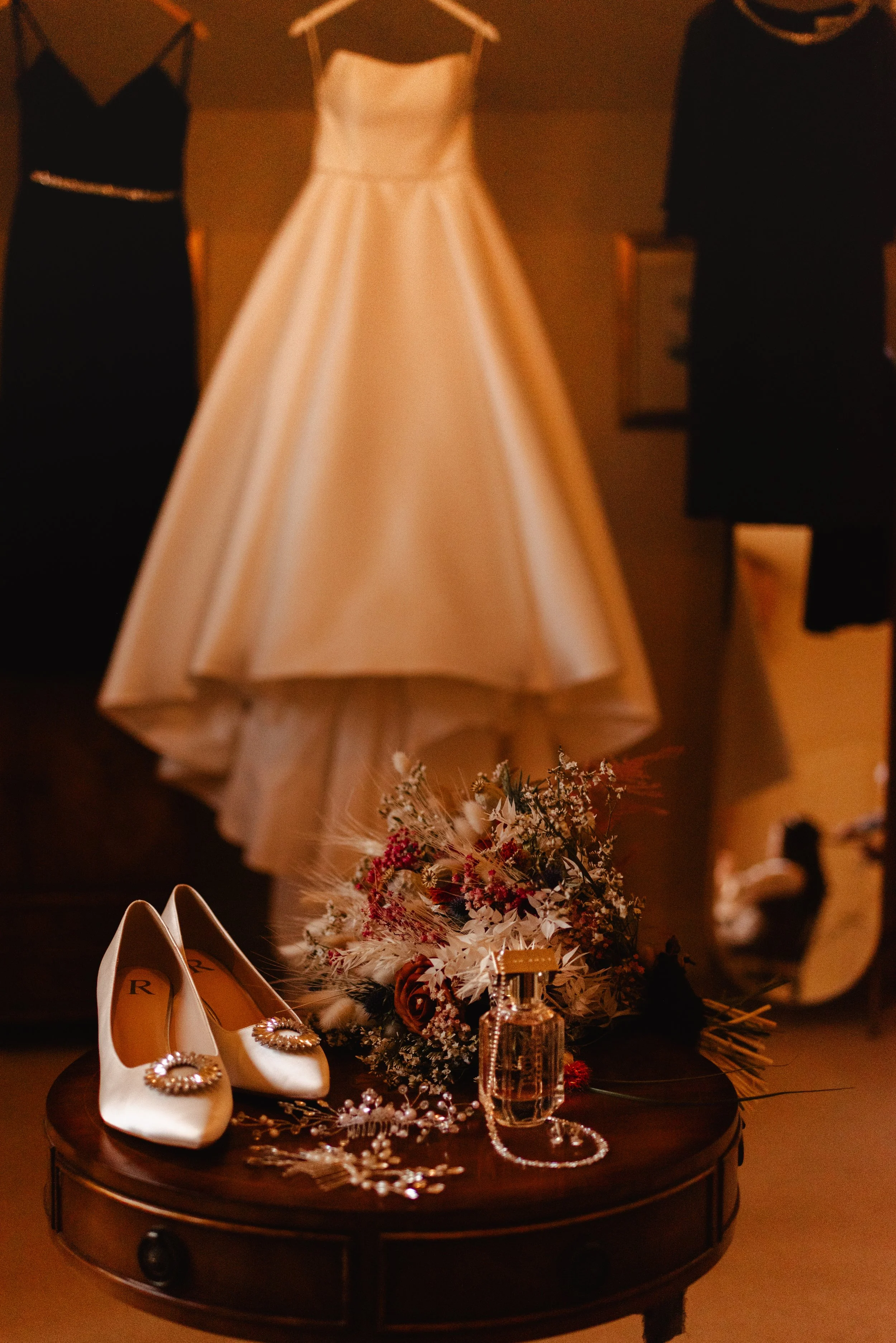 Wedding dress hanging, with black and dark navy dresses in the background. Shoes, a floral bouquet, jewelry, and perfume on a round wooden table in front.