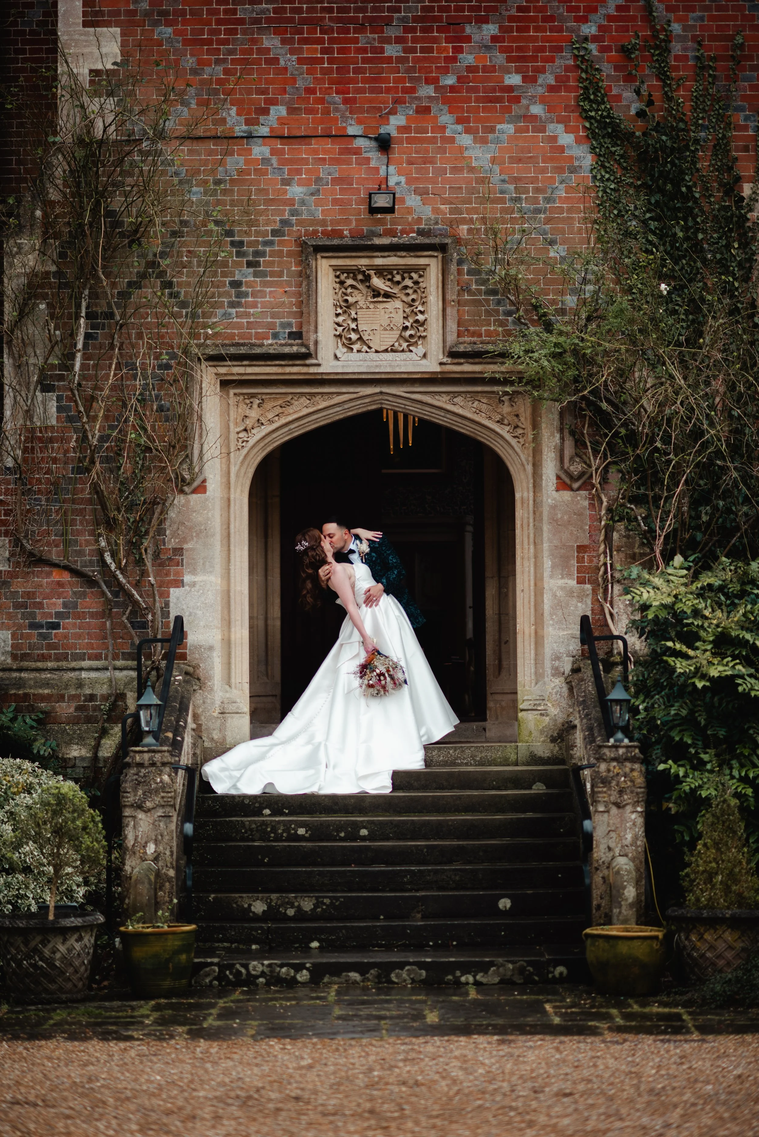 A bride and groom kissing on the steps of an old brick building at their wedding. The bride is wearing a white wedding gown and holding a bouquet of flowers. The groom is dressed in a dark suit. The setting is outdoors with plants and old-fashioned l