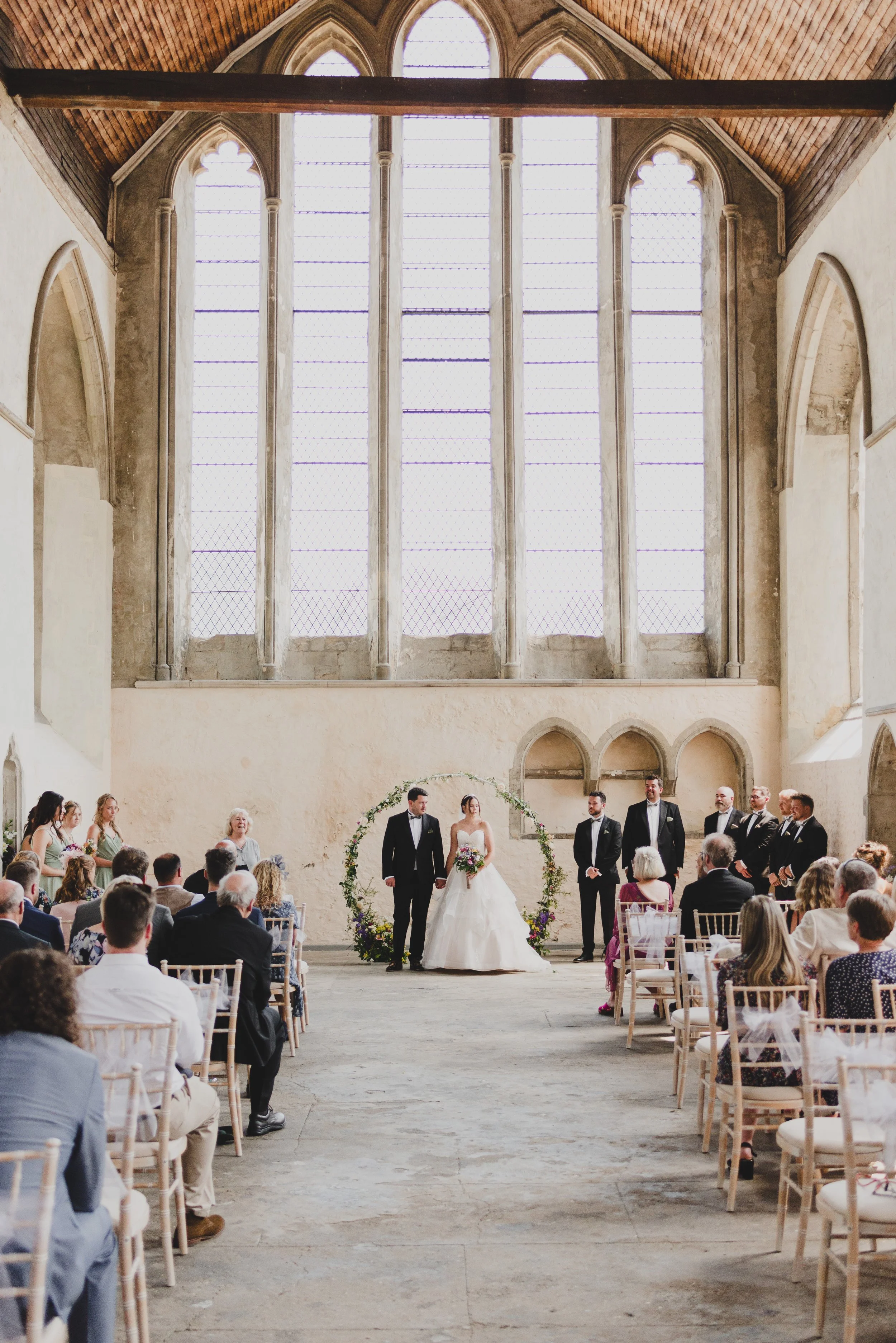 Wedding ceremony in a historic church with high arched window and wooden ceiling. Bride and groom stand under a floral arch with officiants and attendants nearby, guests seated facing them.