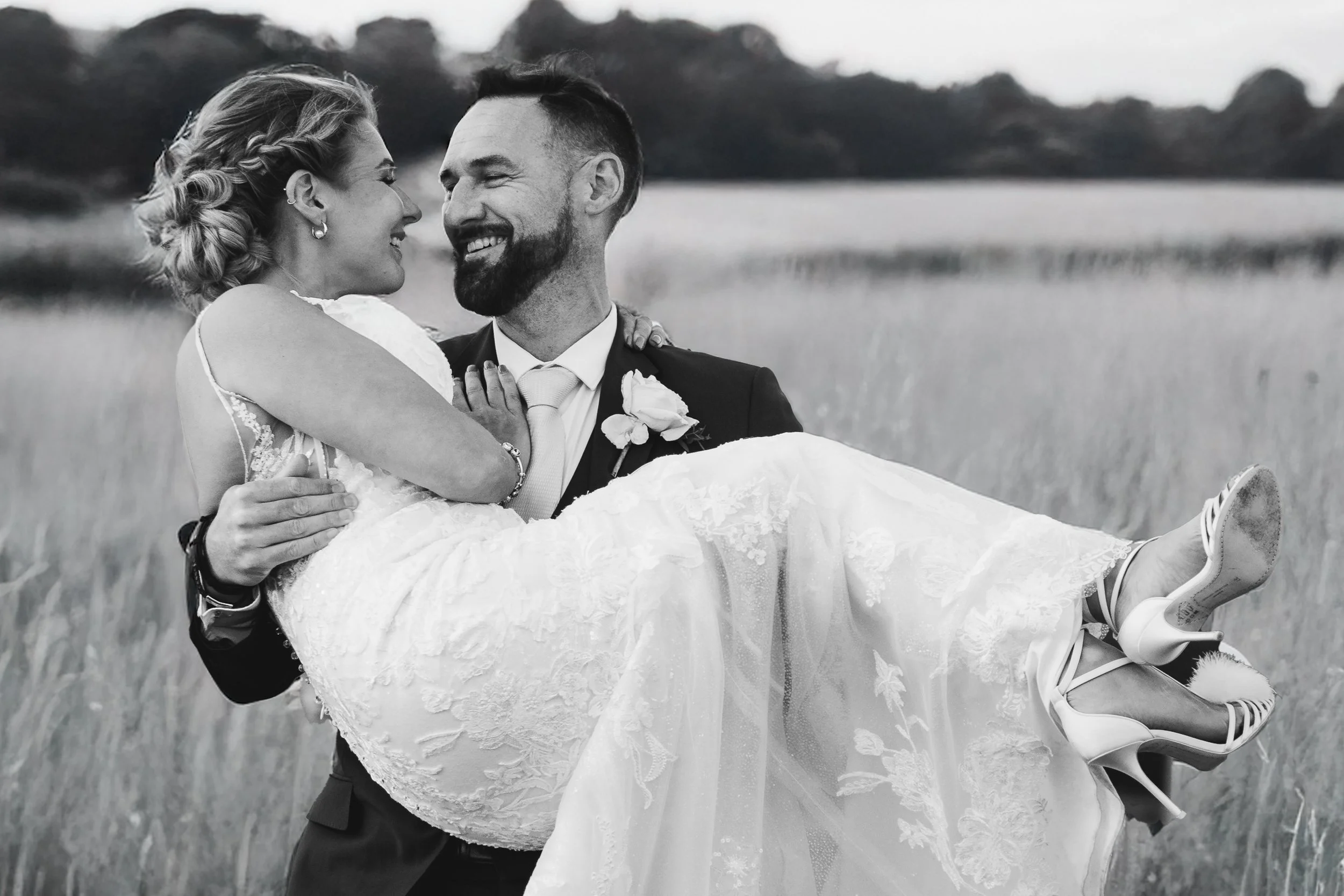 A black and white photo of a groom carrying a bride in a field, both smiling and looking at each other, with mountains in the background.