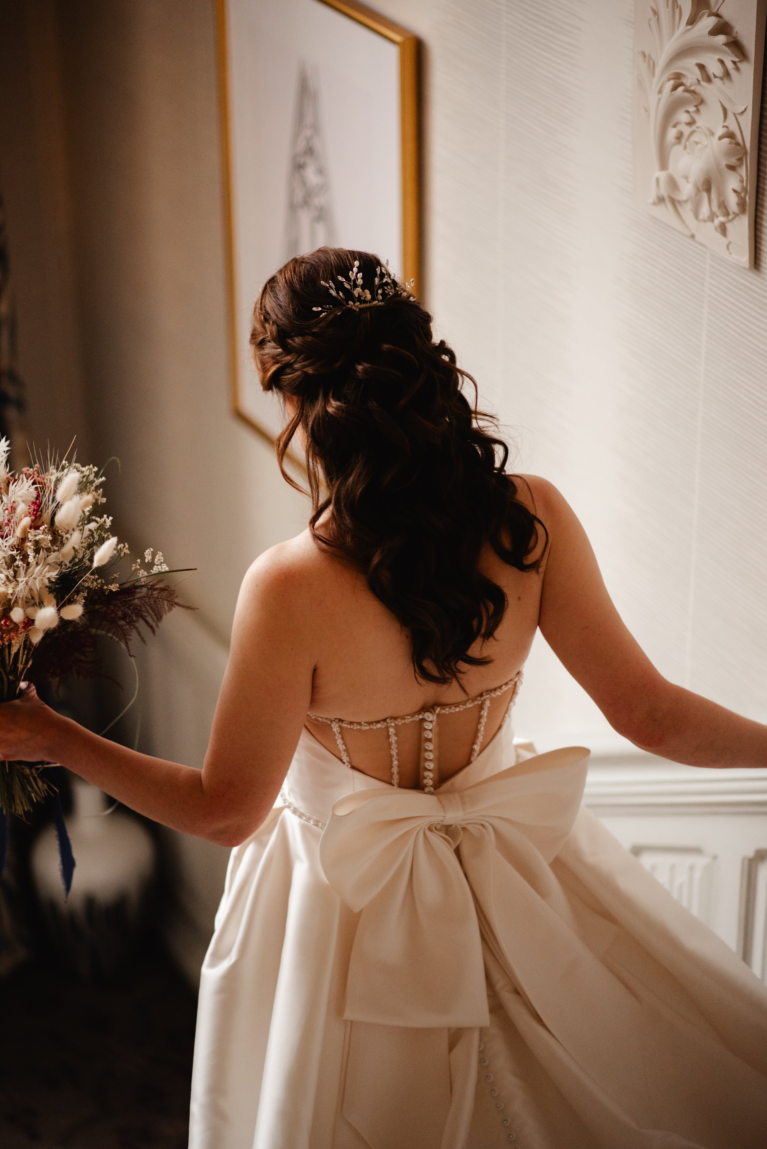 A woman with long, dark, curly hair styled in loose waves and a delicate hairpiece, wearing an elegant, backless wedding gown with pearl embellishments and a large bow at the waist, holding a bouquet of dried flowers, standing in a softly lit room wi