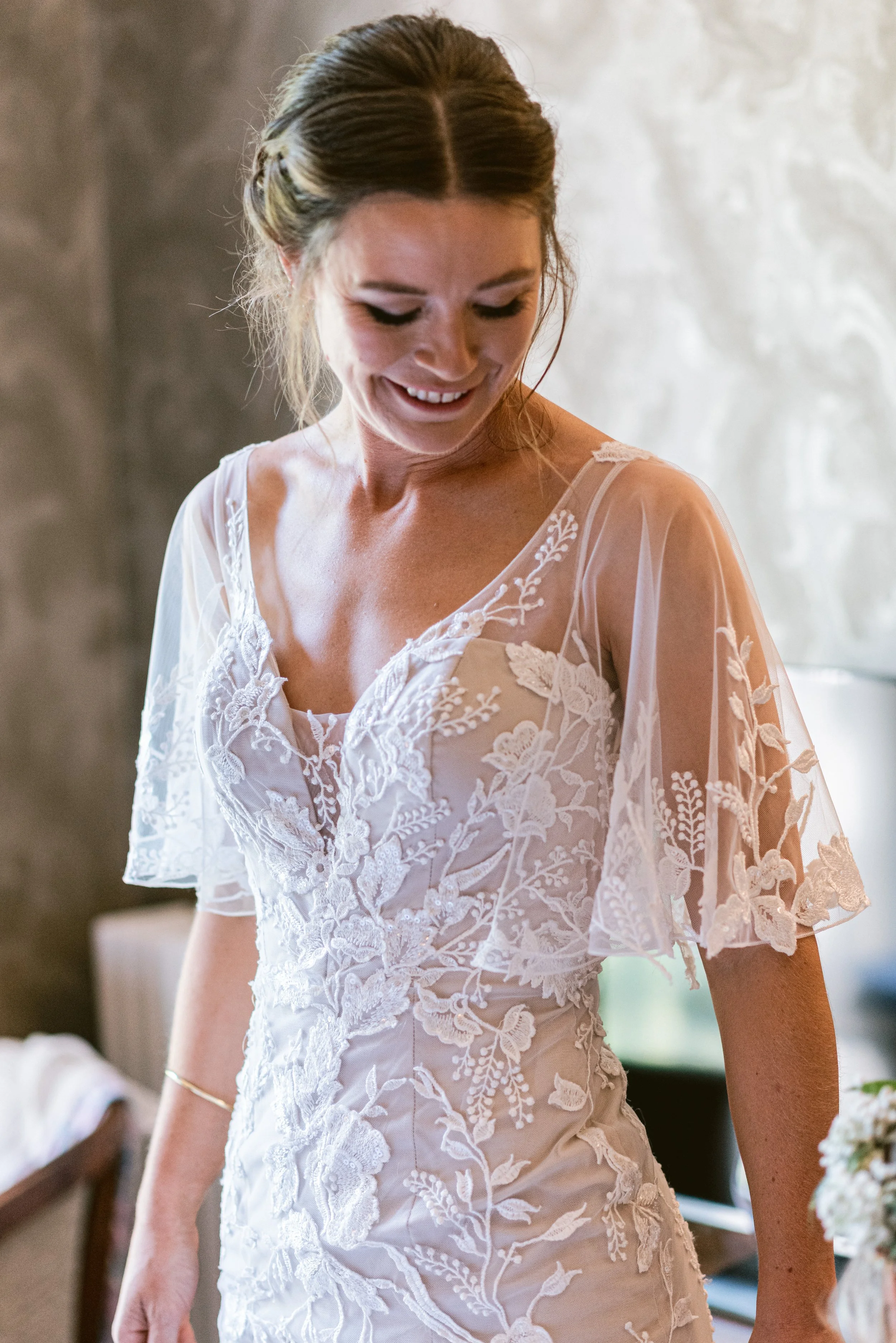 A woman smiling while wearing a white wedding dress with lace embroidery and sheer sleeves.