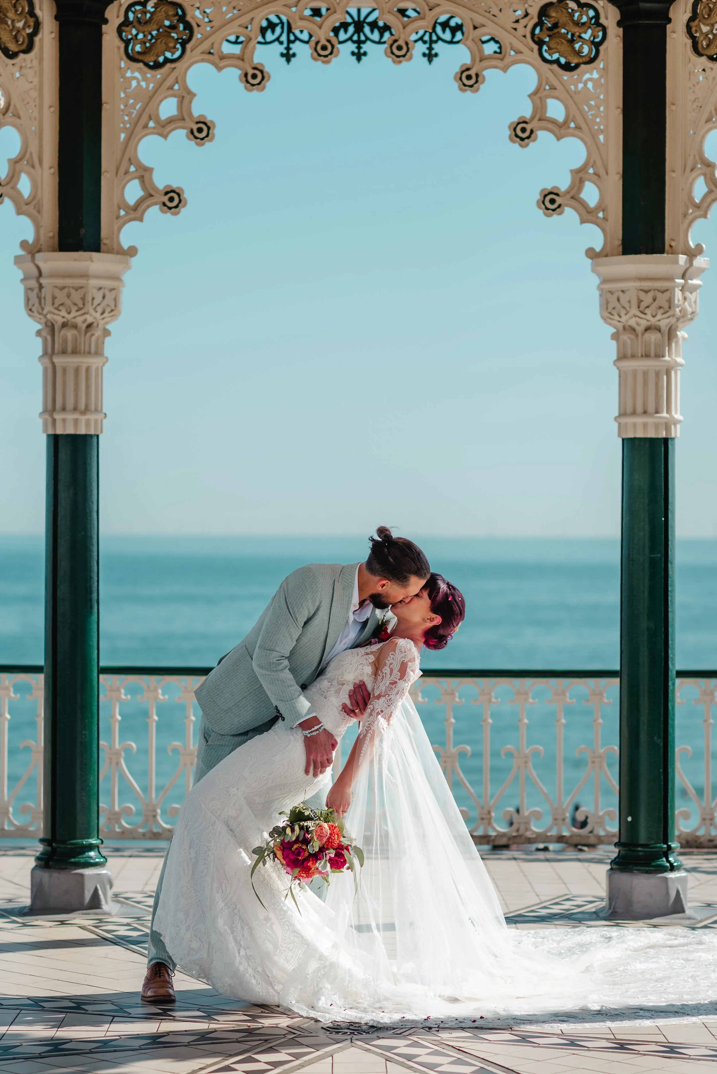 A bride and groom sharing a kiss on a balcony overlooking the ocean, with ornate architectural details framing the scene, and the bride holding a bouquet of flowers.