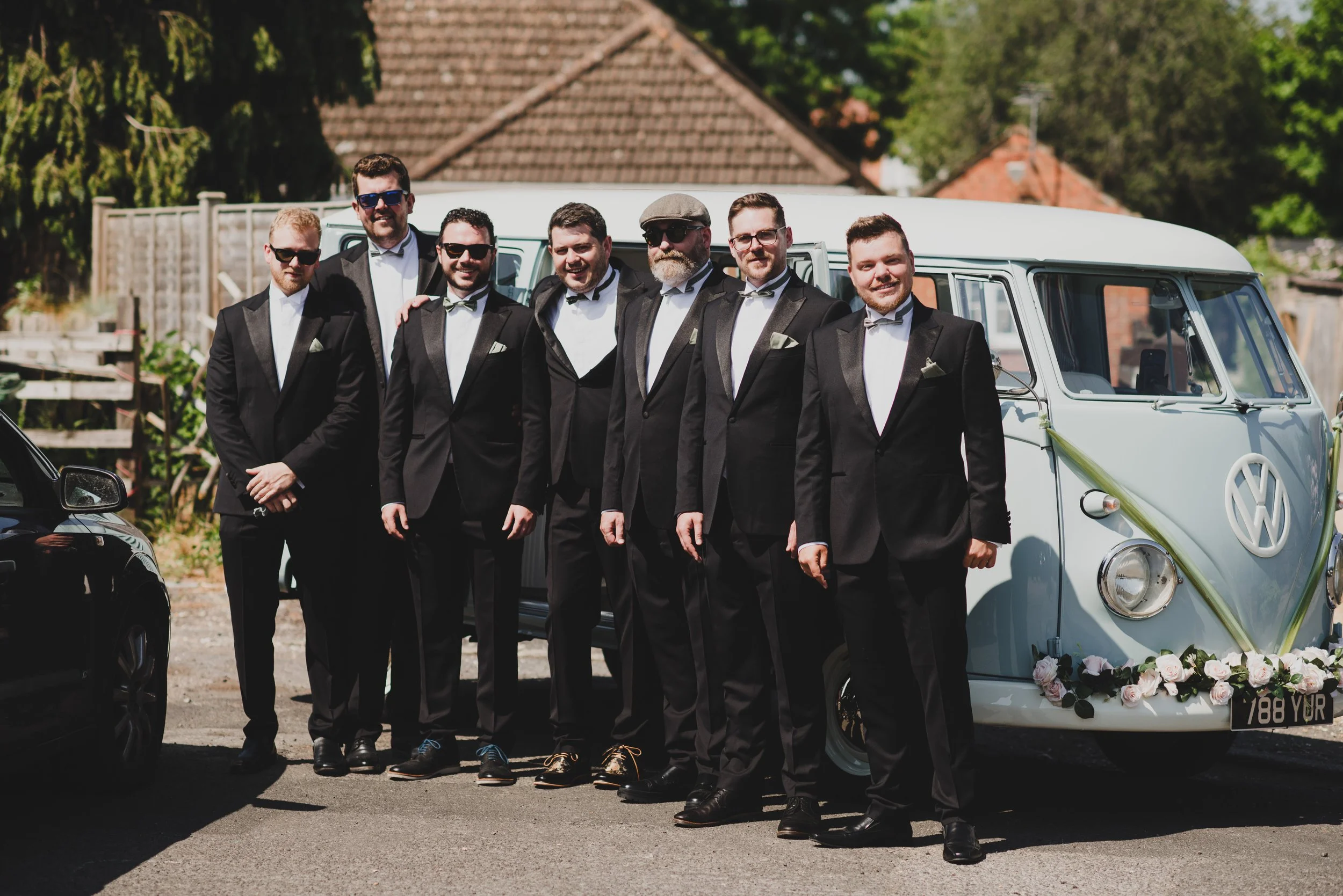 A group of seven men in formal tuxedos with bow ties standing in front of a vintage Volkswagen van decorated with flowers for a wedding or special occasion.