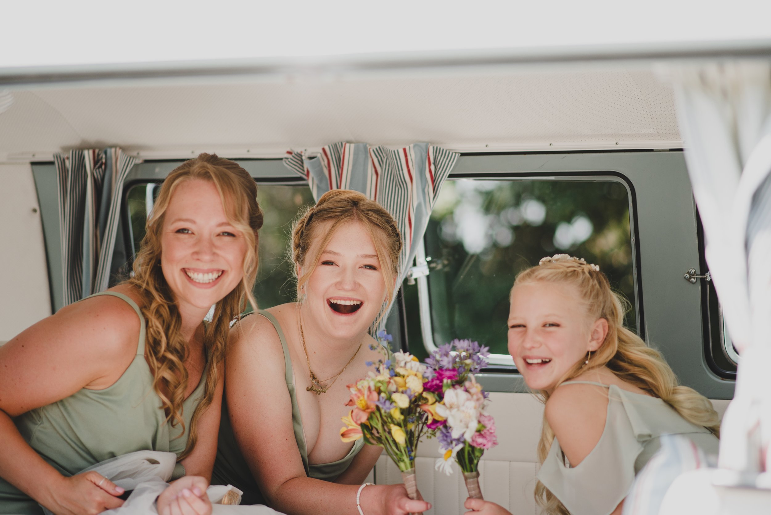 Three women, smiling, inside a vehicle with striped curtains, holding bouquets of flowers.