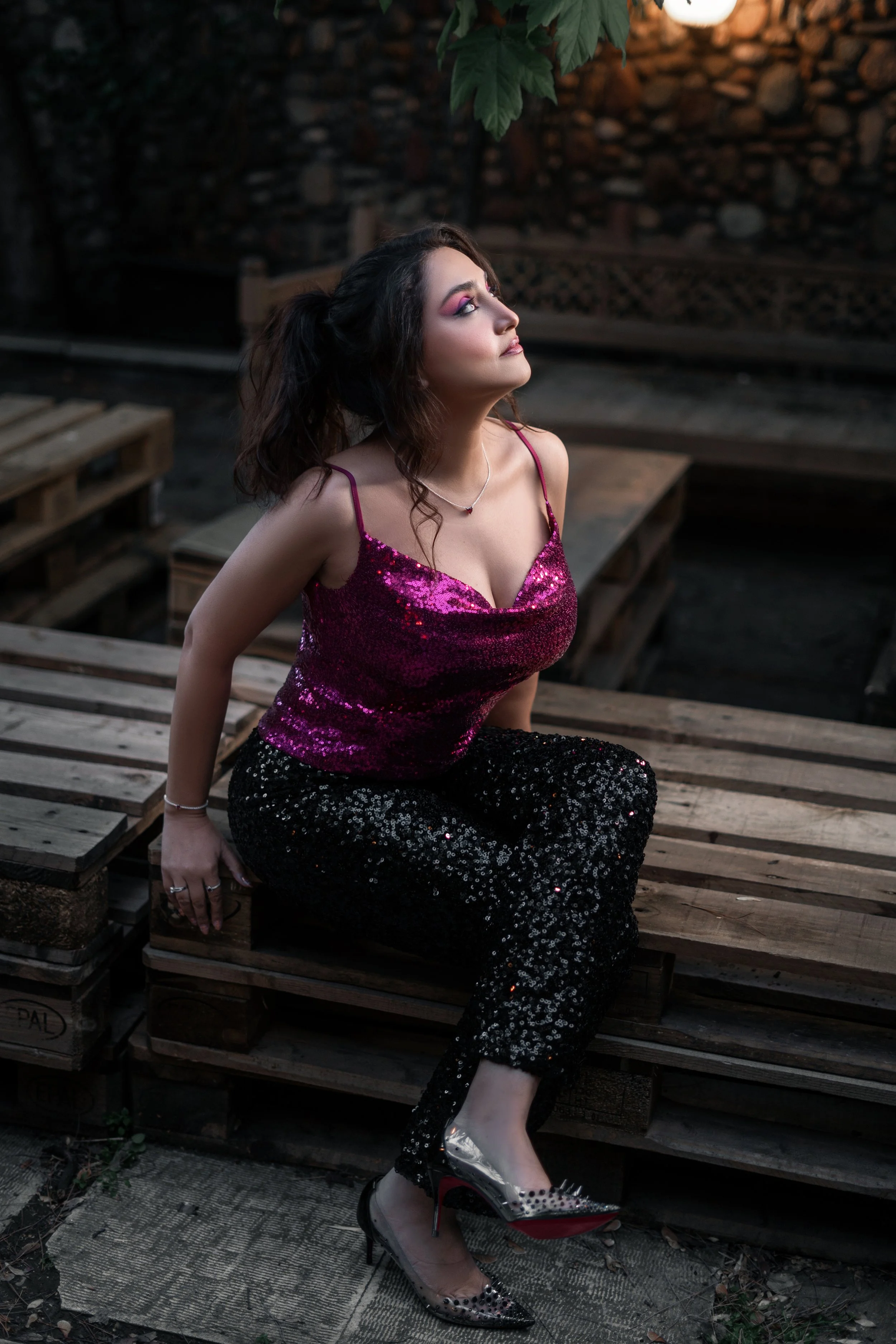 A woman with dark wavy hair wearing a sparkly pink top, black sequin pants, and clear high heels with red soles, sitting on wooden pallets outdoors.