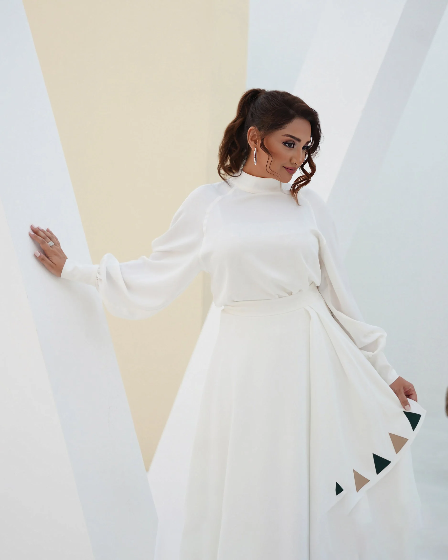 A woman with dark hair in loose curls, wearing a long white dress with a high neckline and long puffed sleeves, standing against a white and soft yellow background.