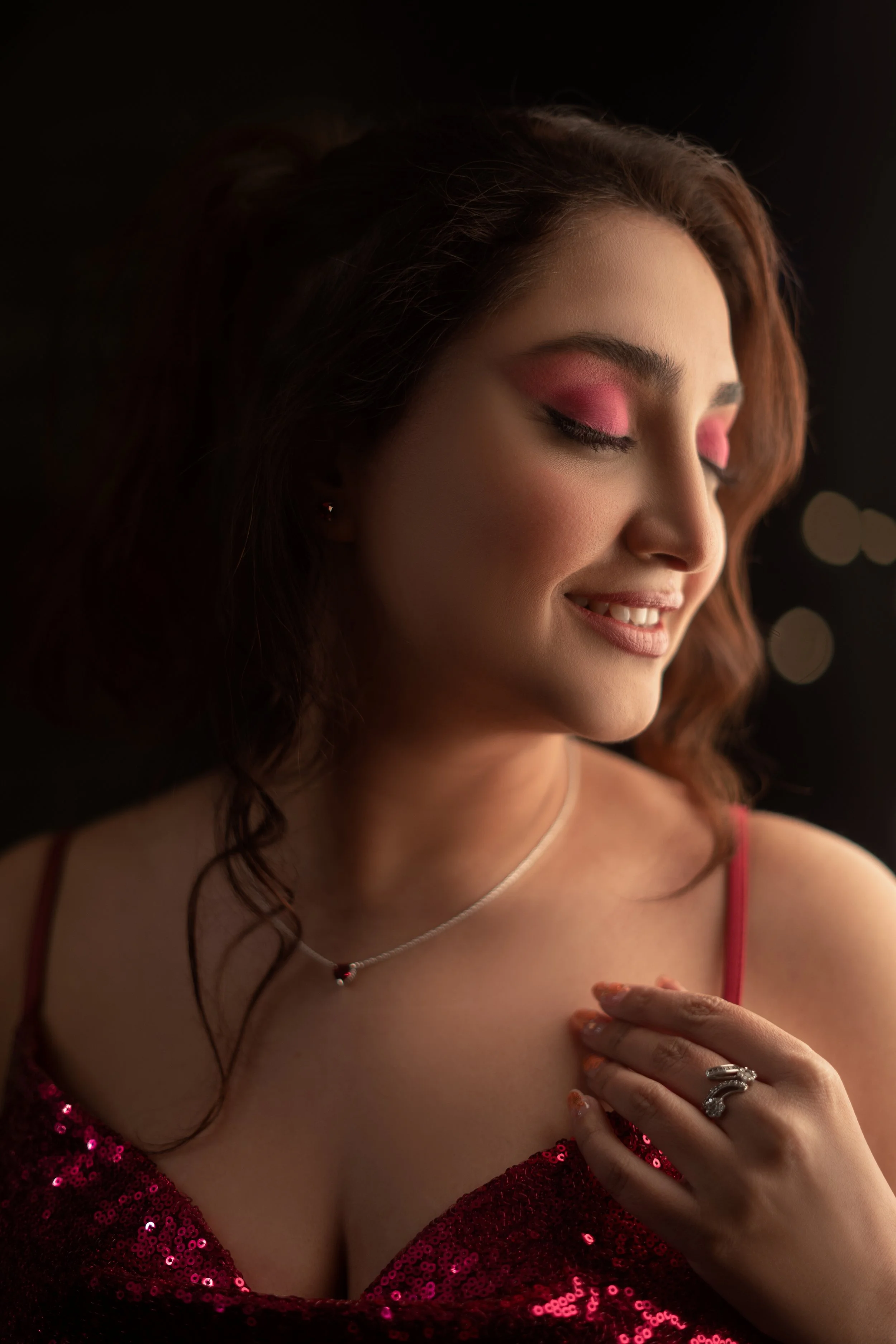 A woman with red hair, pink eyeshadow, and a sparkling red dress, smiling with her eyes closed.