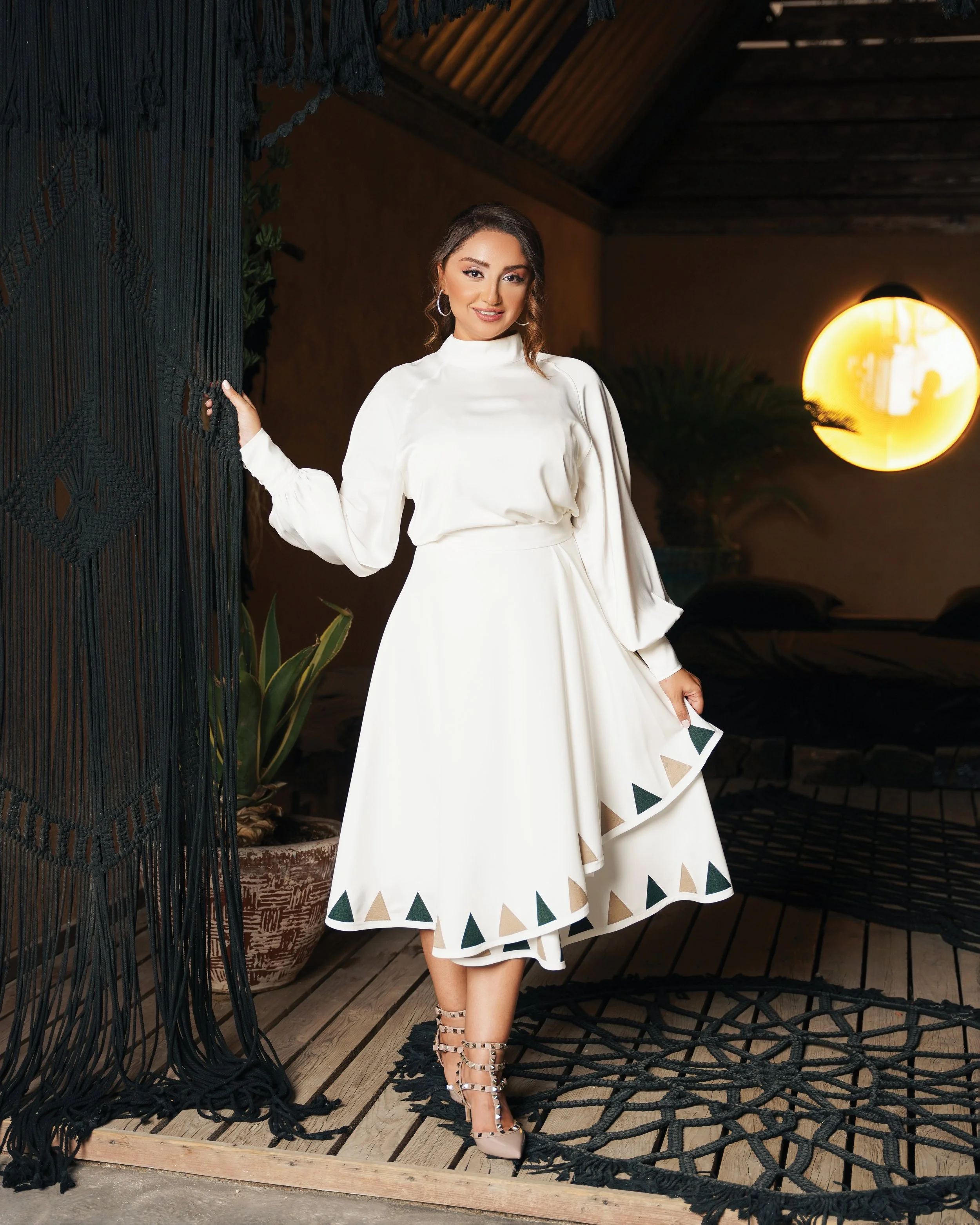 A woman with brown hair styled in loose waves, wearing a white long-sleeve dress with a high neckline, standing indoors at night. She is holding the curtain with one hand and lifting her skirt slightly with the other, showing beige and black triangul