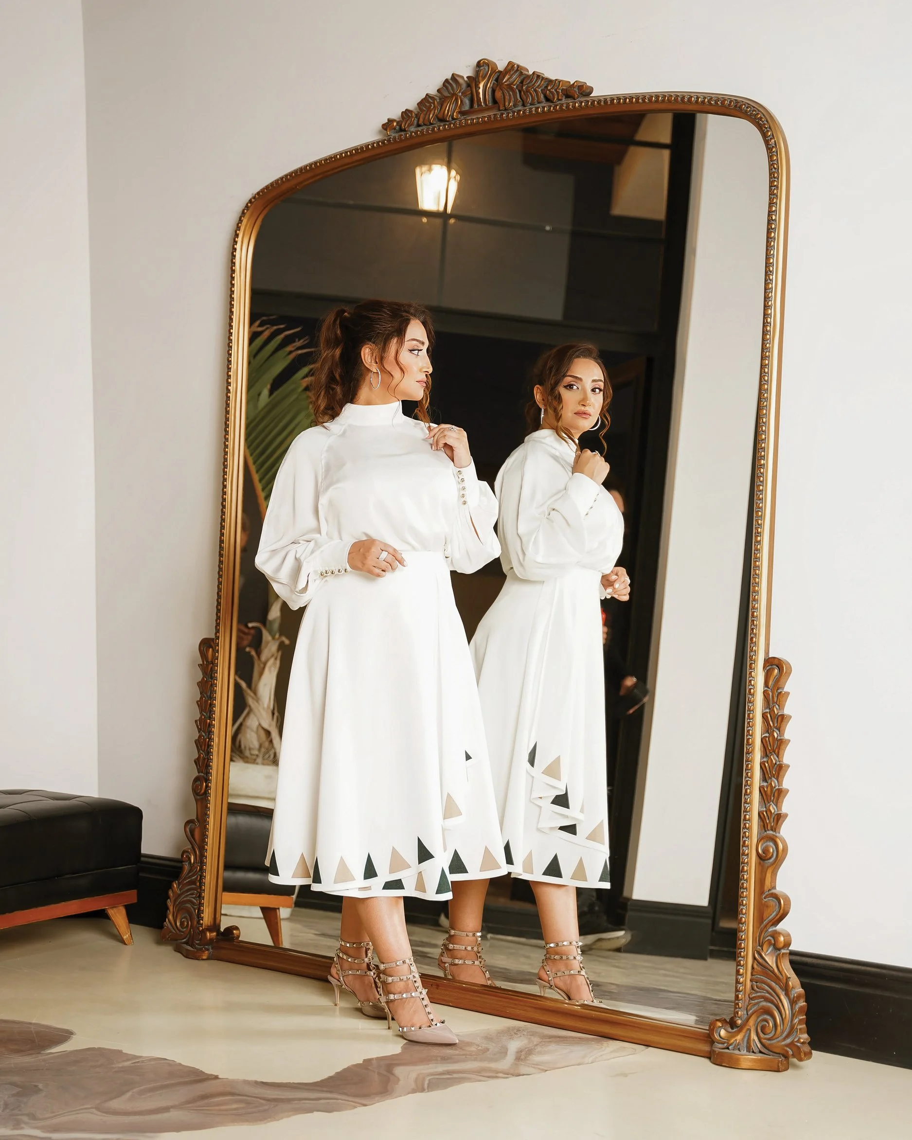A woman in a white dress with triangle cutouts on the hem stands in front of a large ornate mirror, adjusting her sleeve. She is wearing high heels and has her hair styled in loose waves. The reflection shows her face and part of her profile.