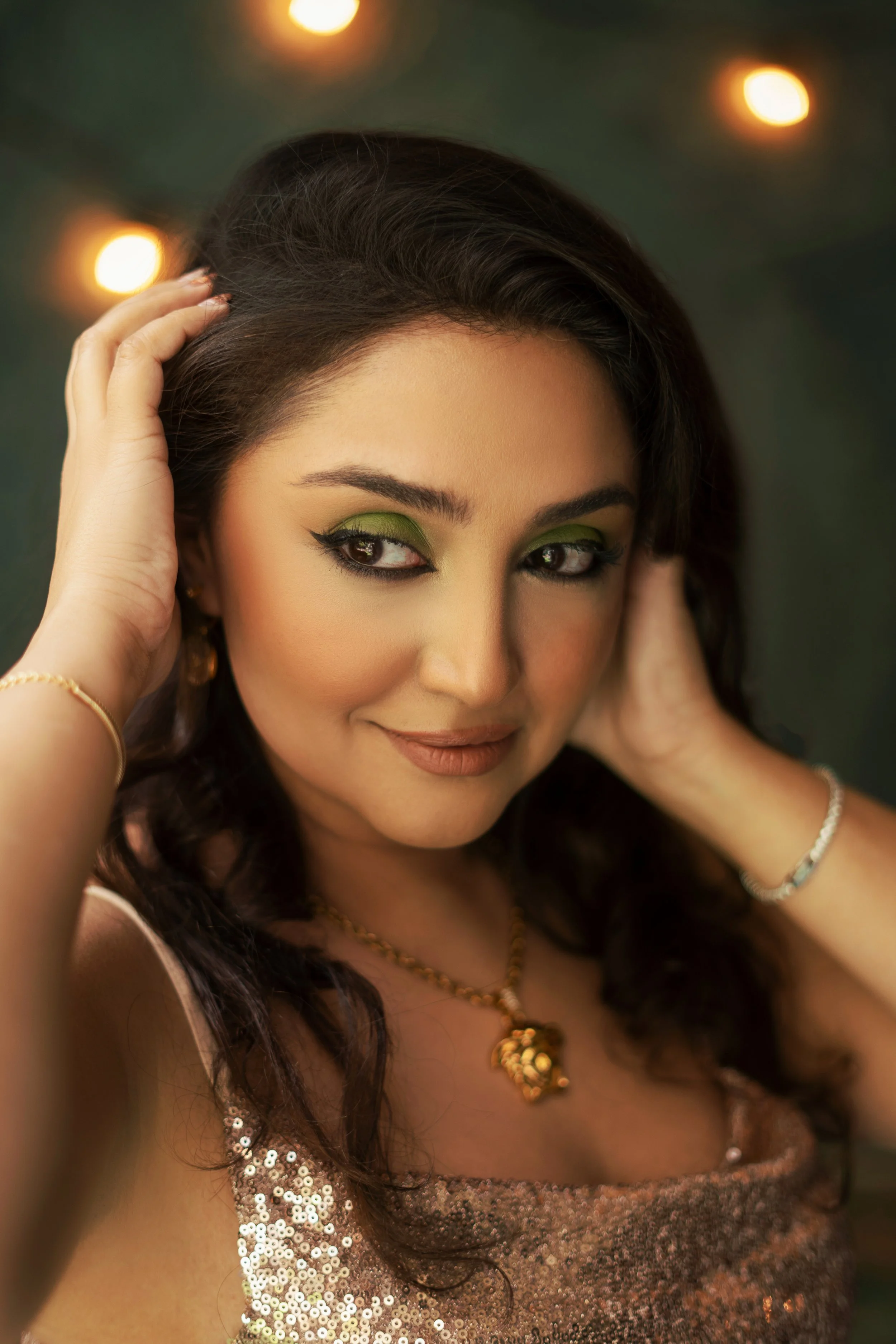 A woman with dark hair and green eyeshadow, wearing a sparkling gold top and gold jewelry, looks at the camera while touching her hair with both hands against a blurred background with lights.