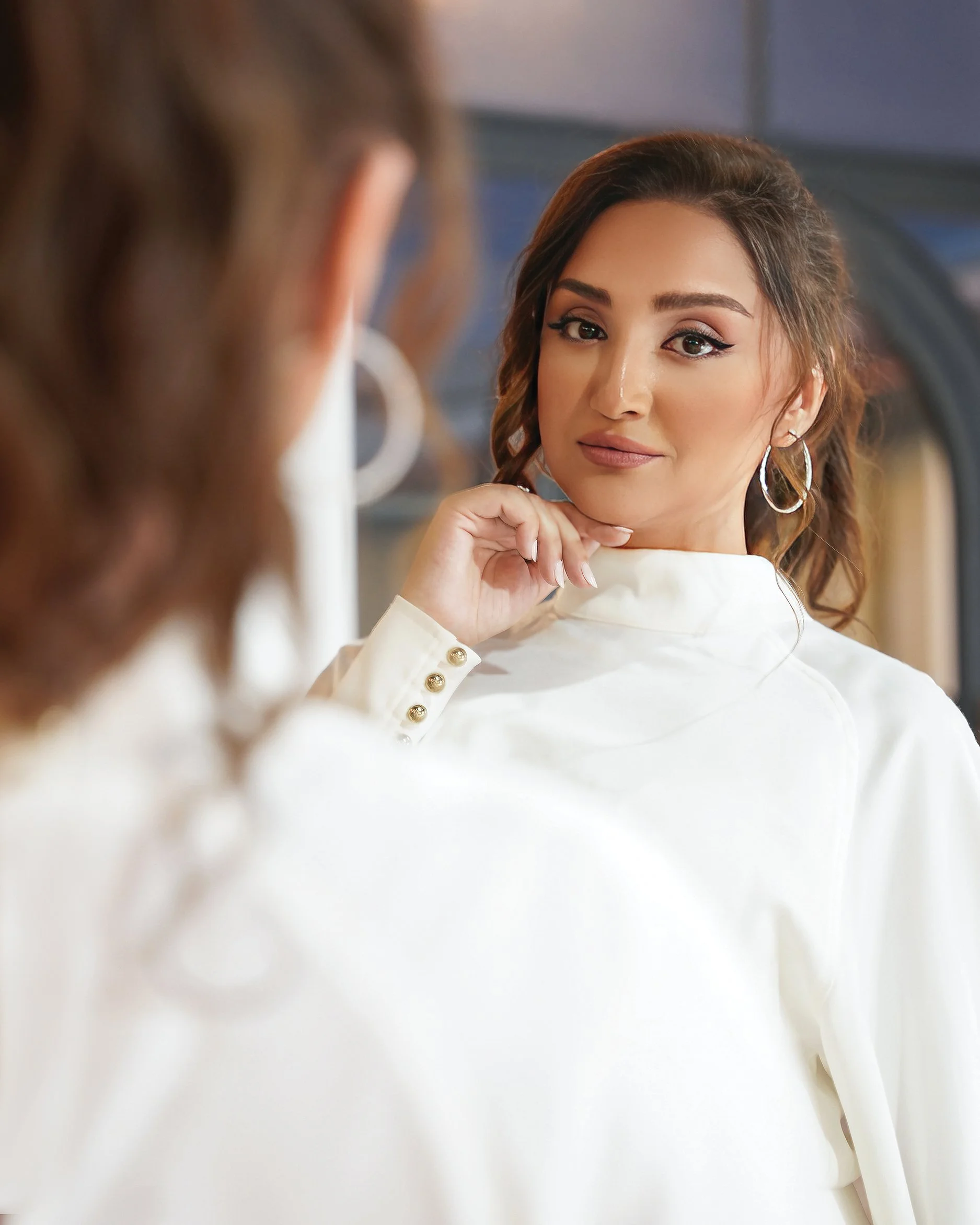 A woman with brown hair styled in loose curls, wearing a white blouse and hoop earrings, looks at herself in a mirror with a confident expression.