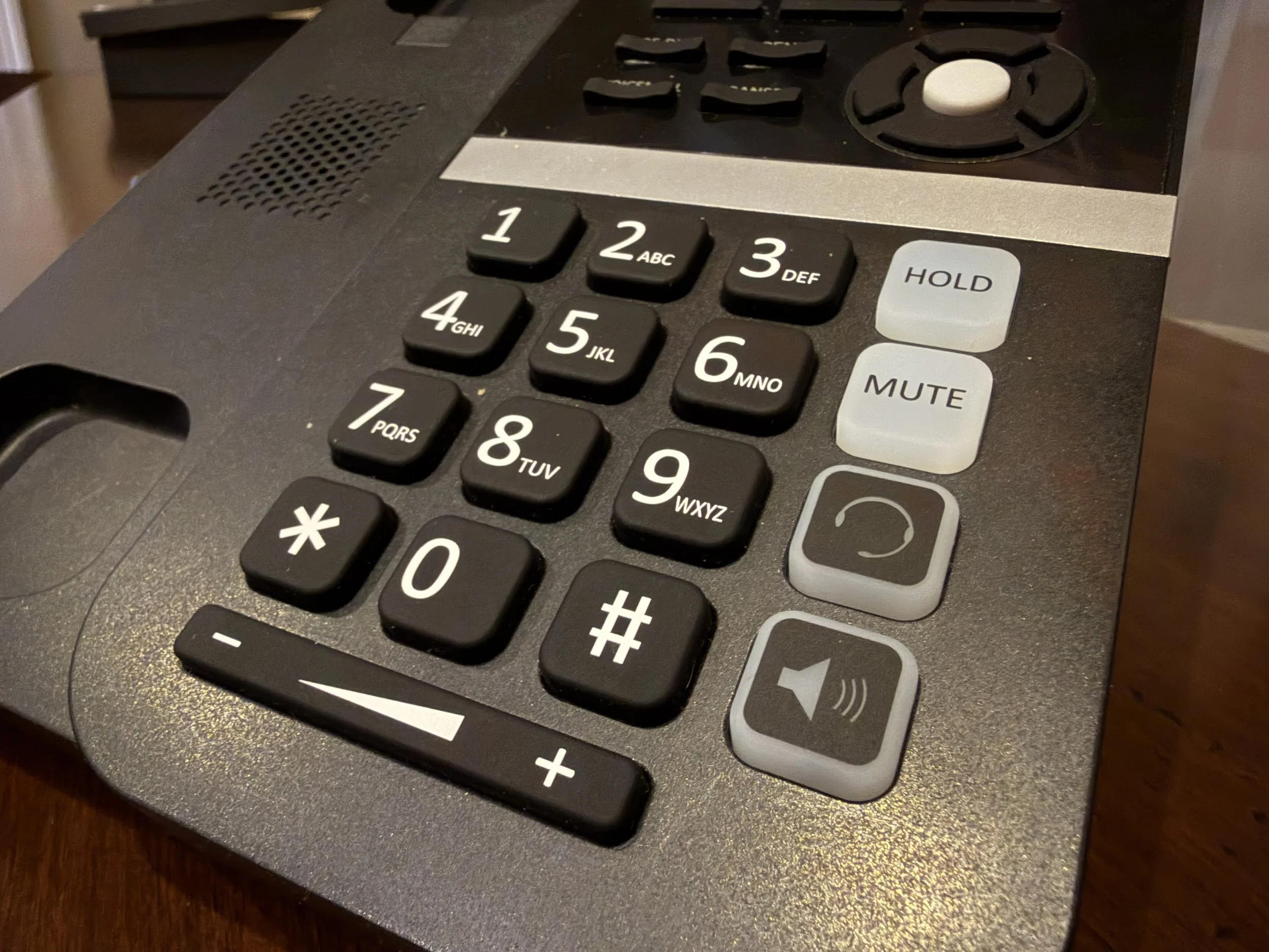Close-up of a office landline phone keypad with numbers 0-9, *, #, and additional buttons for hold, mute, and speakerphone on a wooden surface.