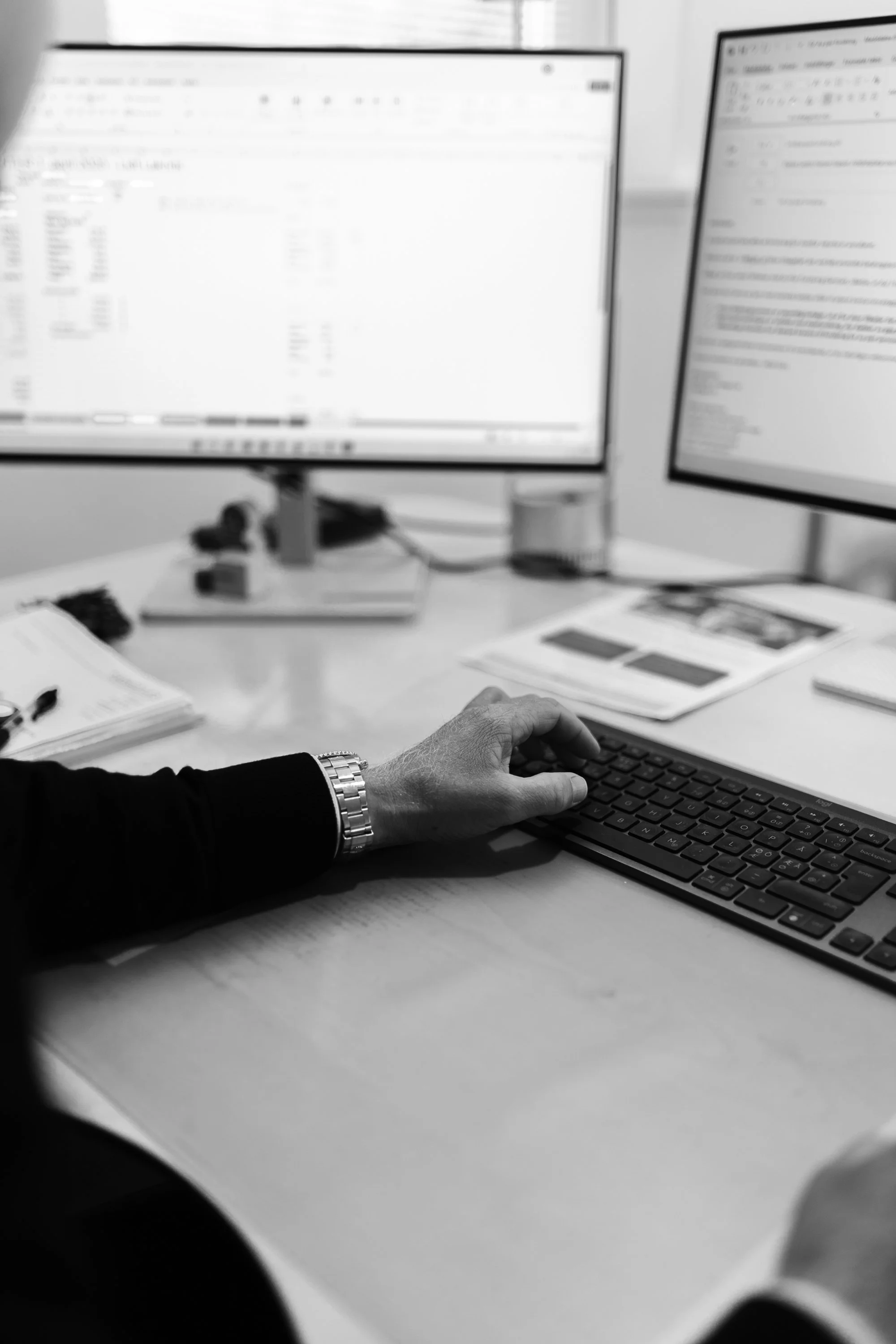 Hænder skriver på tastatur foran to computerskærme, kontorarbejde med forsikringsløsninger.