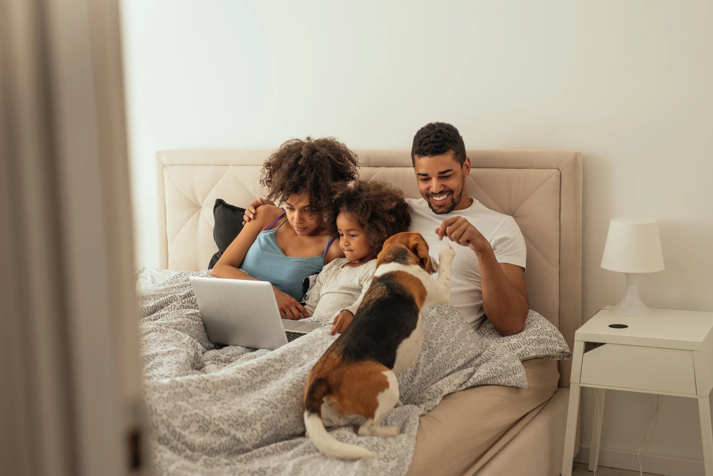 Family of three, two children and a father, sitting on a bed and looking at a laptop with a dog reaching up to them.