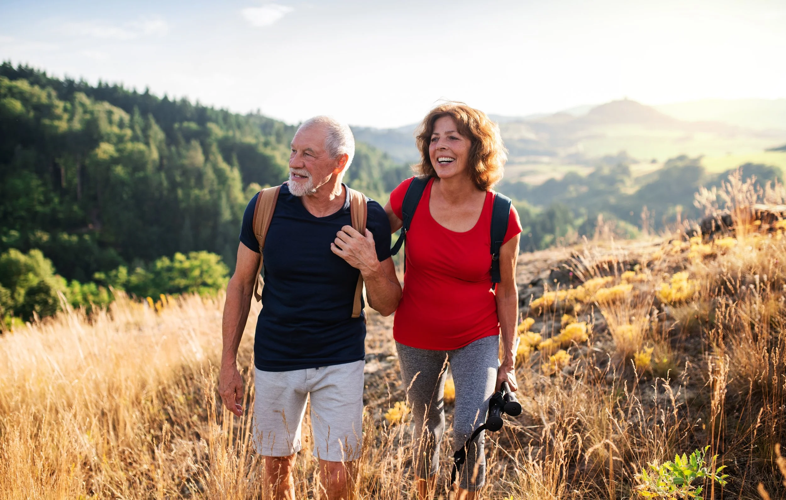 A smiling senior couple hiking on a trail through a grassy field with mountains and trees in the background.