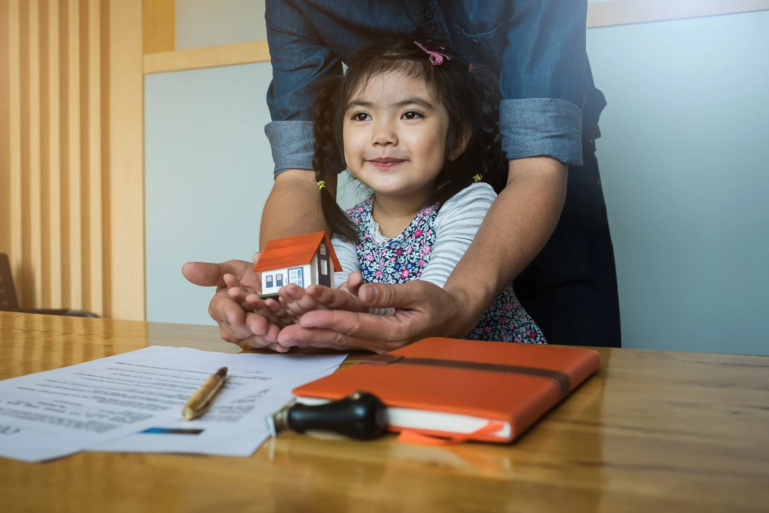 A young girl with pigtails and a floral shirt holding a small model house with an adult helping her. The scene is at a table with papers, a pen, a notebook, and a car key.