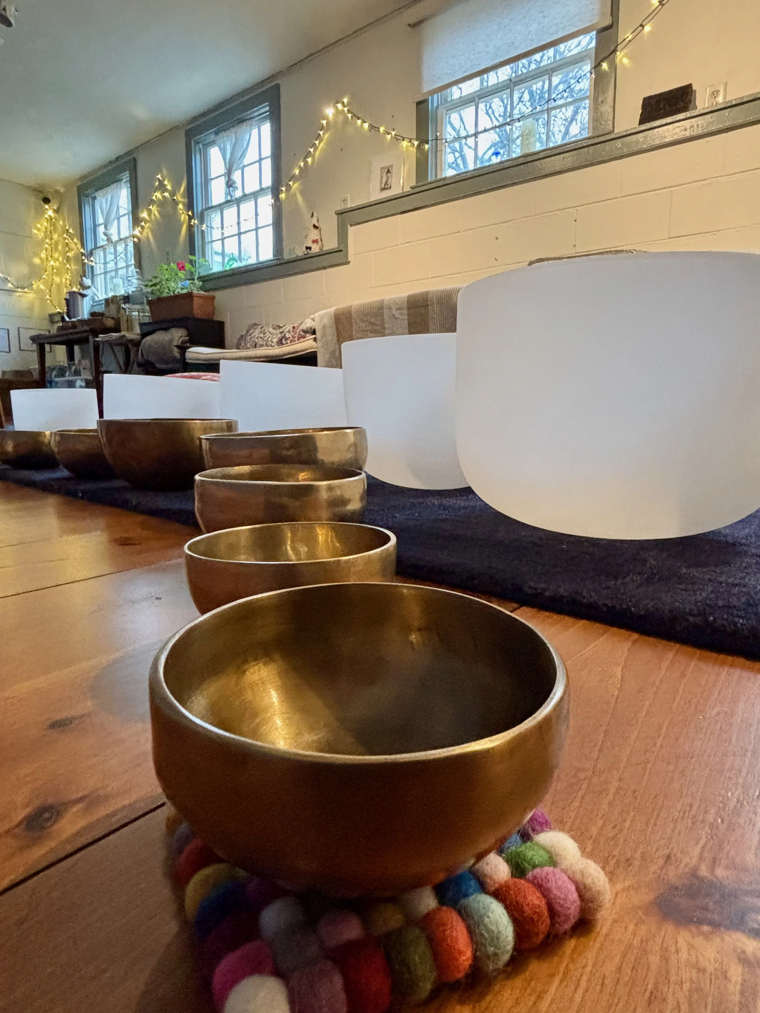 A row of brass singing bowls on a colorful felt mat, placed on a wooden floor in a room with string lights on the wall, windows, and various objects on a table in the background.