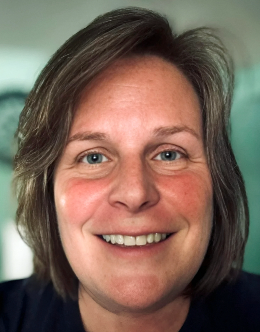 Close-up of a smiling woman with shoulder-length brown hair and blue eyes, wearing a dark top, indoors with a blurred green background.