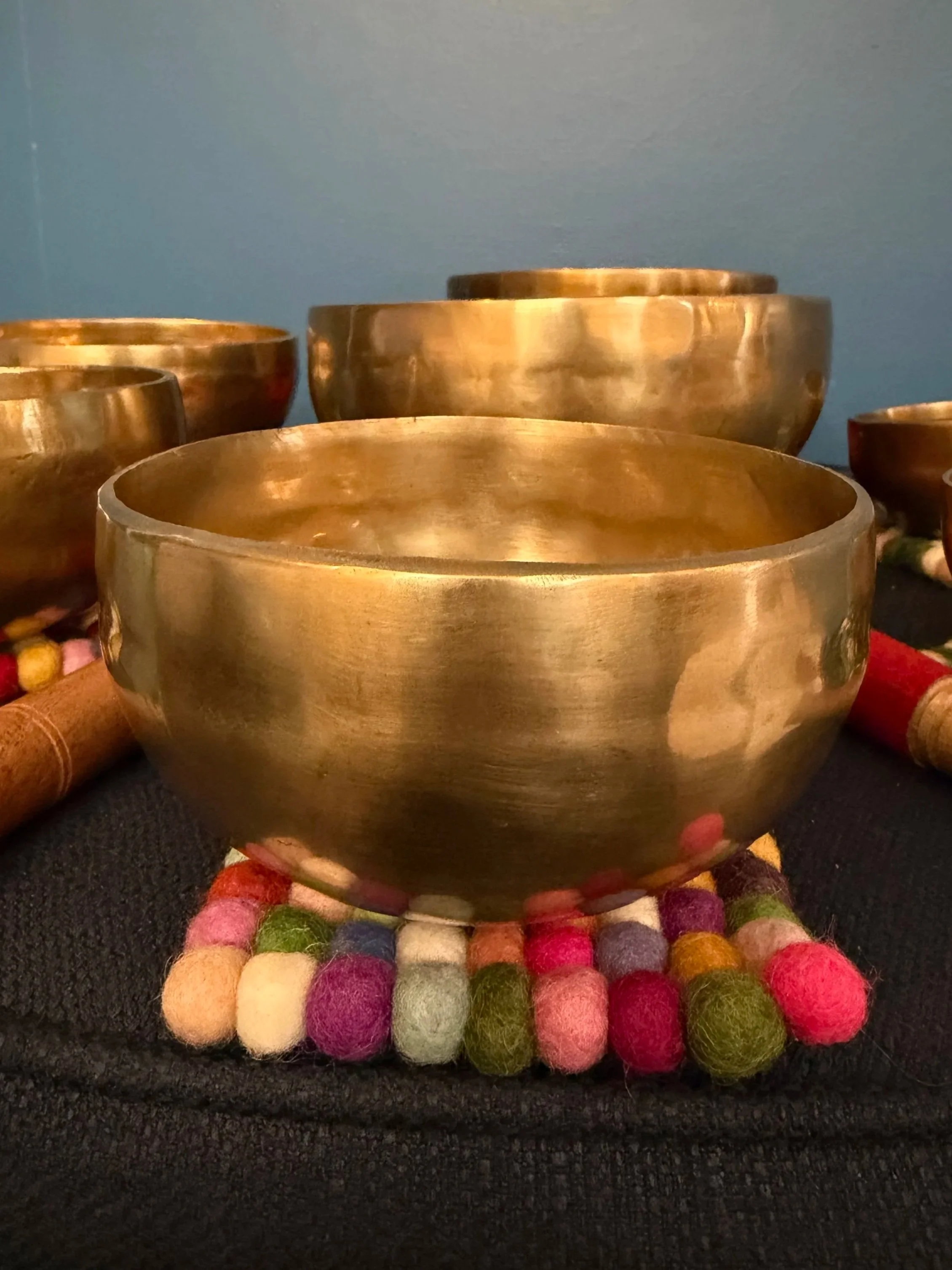 Several brass singing bowls placed on a colorful felted wool mat with a black textured surface background.