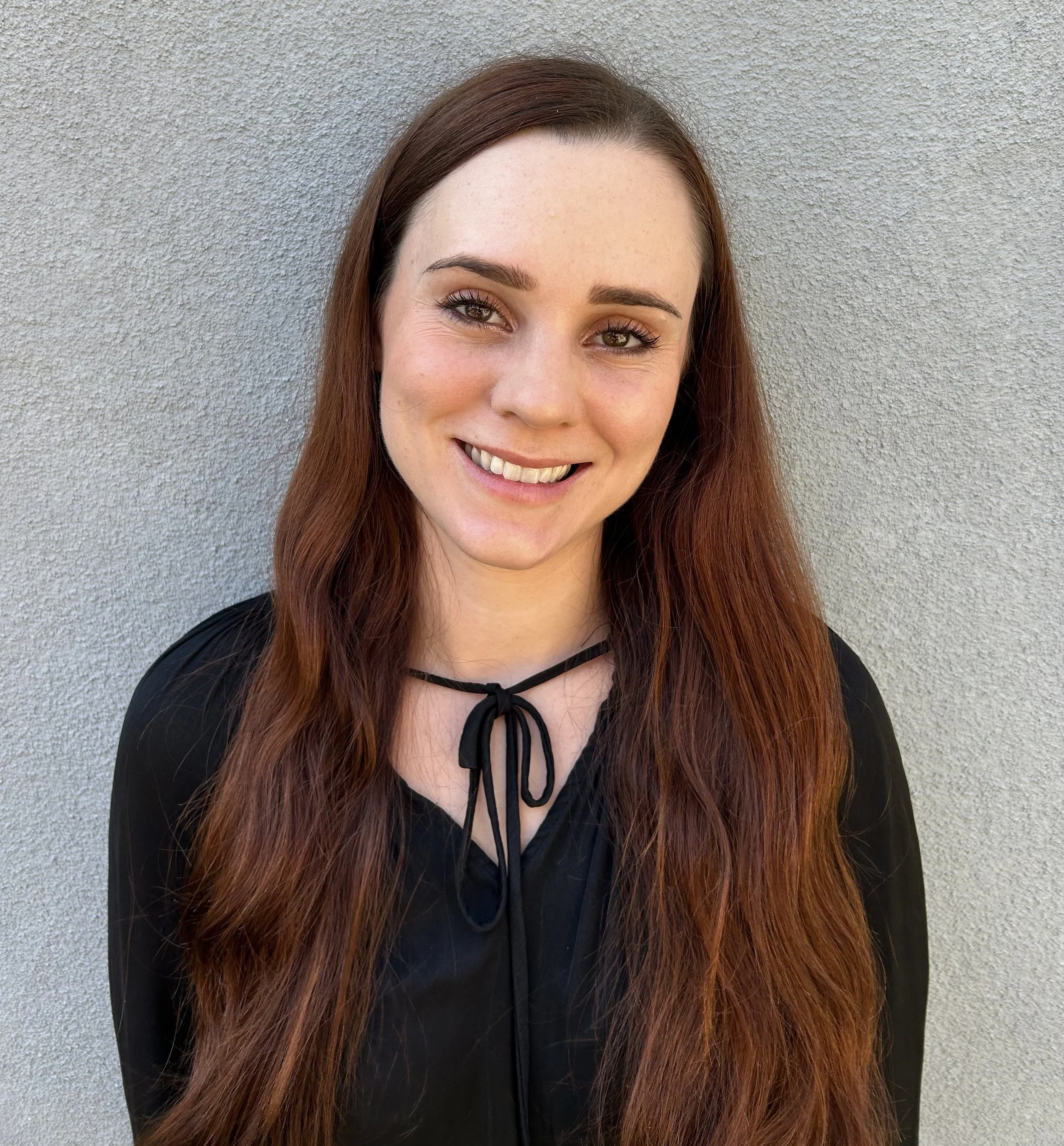 Close-up image of a young woman with long, wavy auburn hair, smiling, standing in front of a textured gray wall.