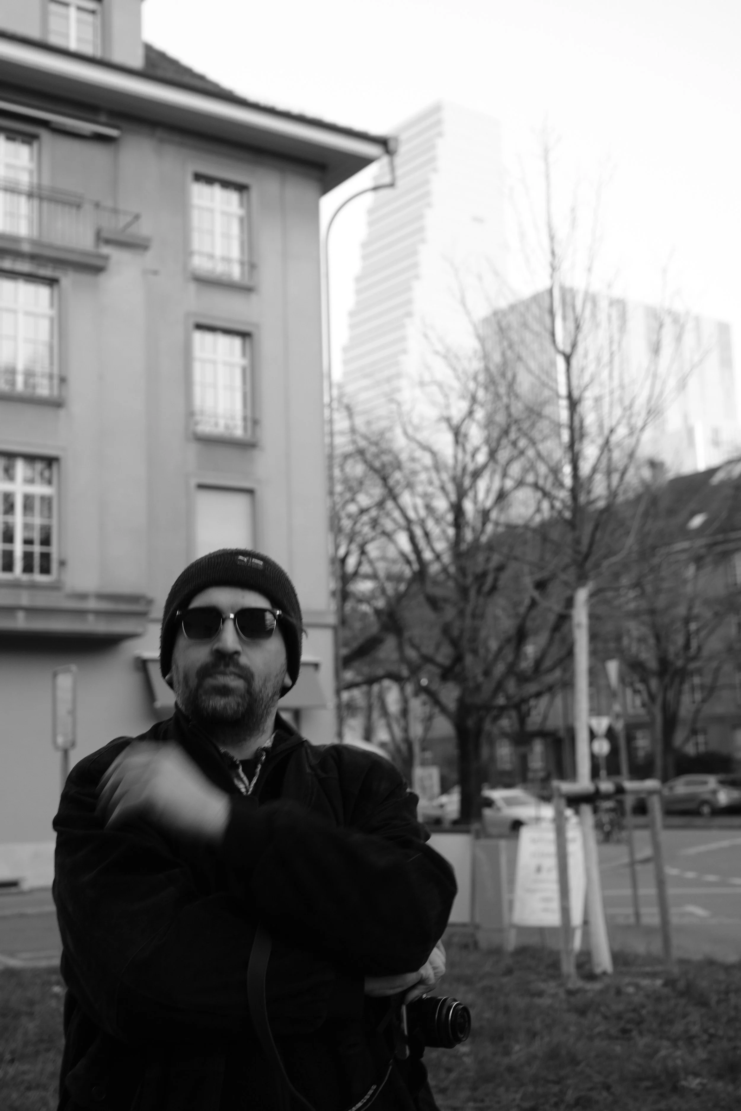 Schwarz-Weiß-Foto eines Mannes mit Sonnenbrille, Mütze und dunkler Jacke, der vor einem mehrstöckigen Gebäude in einer Stadt steht, mit Bäumen und Straßen im Hintergrund.