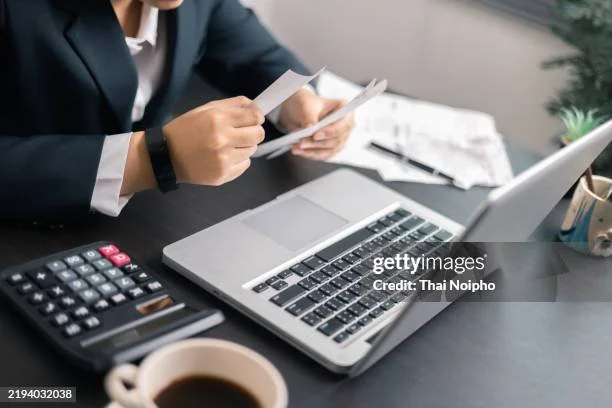A person in business attire sitting at a desk with a laptop, calculator, papers, pen, and a cup of coffee.