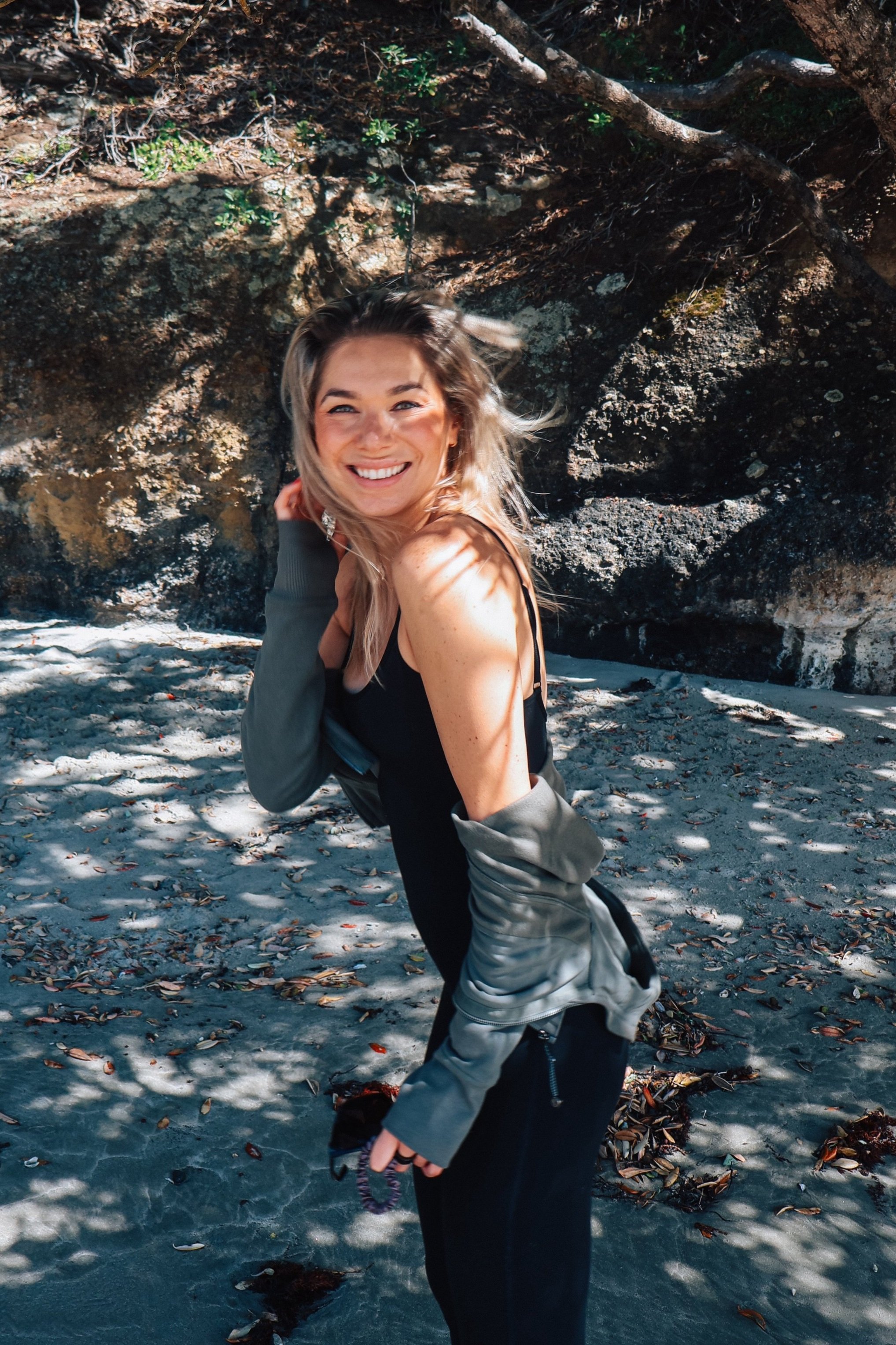 A woman with blonde hair, wearing a black sleeveless top and a green jacket, smiling outdoors on a beach with rocks and fallen leaves, shadowed by trees.