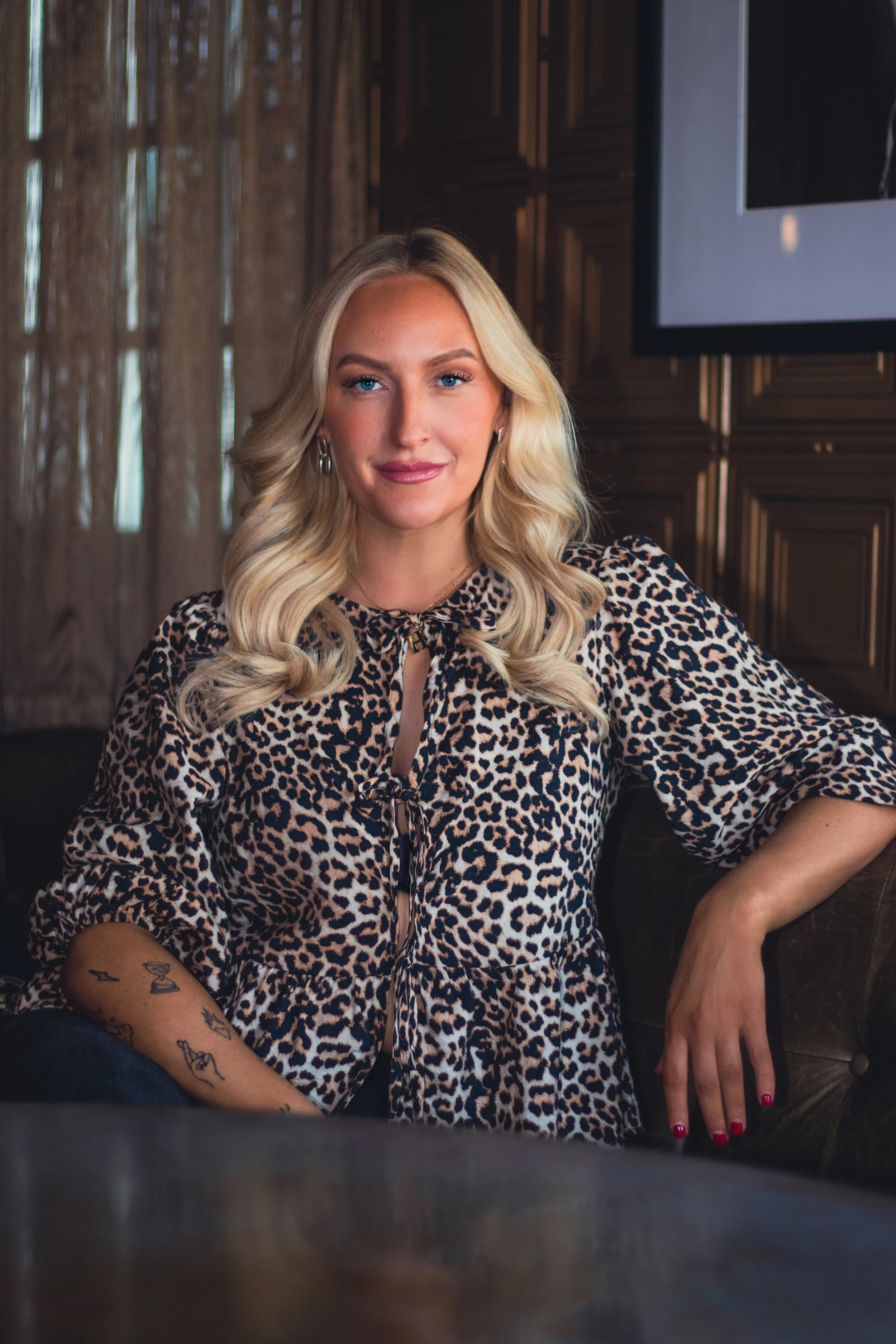 A blonde woman with wavy hair sitting on a couch in a leopard print top, looking at the camera with a slight smile.