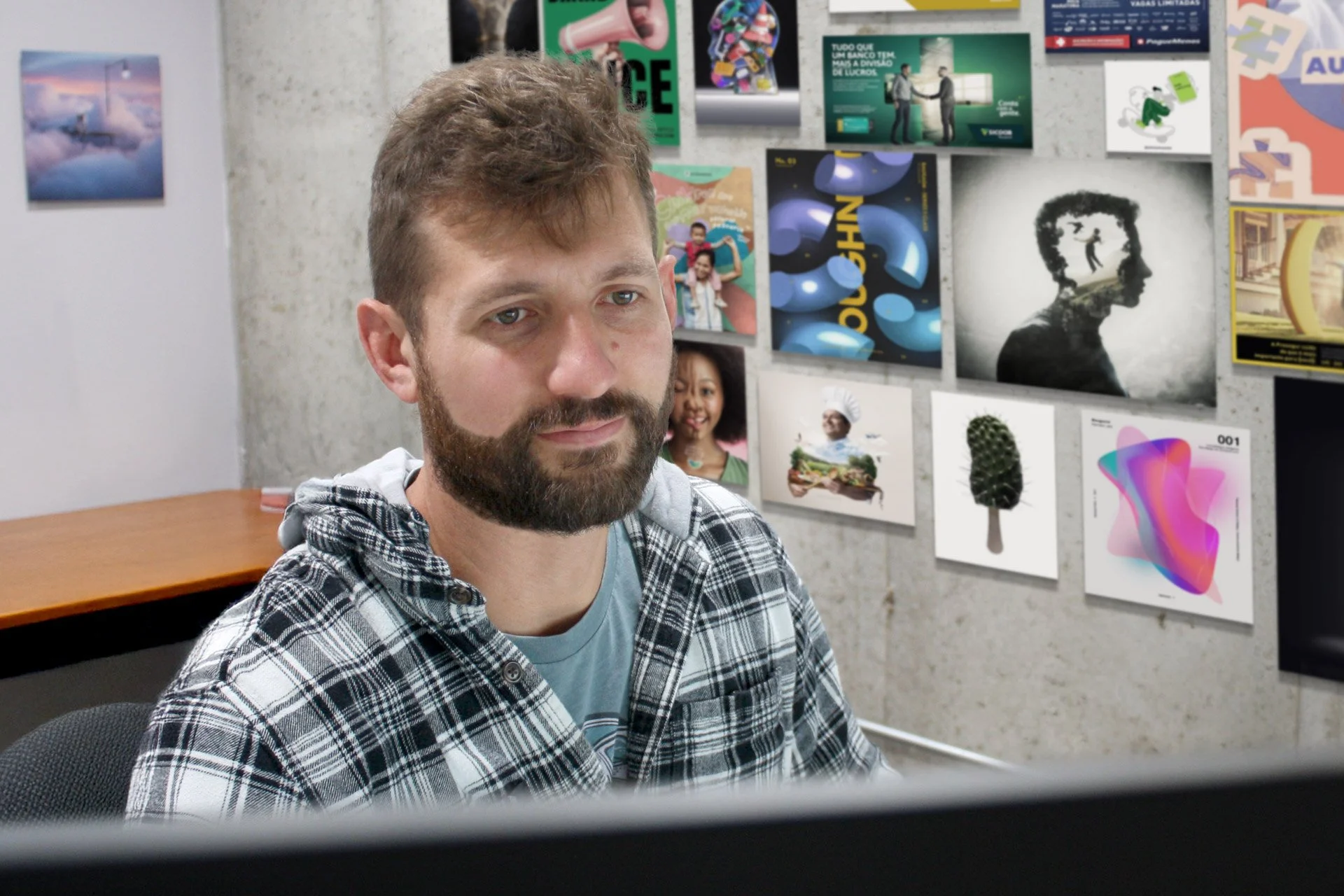 A man with a beard wearing a plaid shirt sitting in front of a computer with a wall of artistic posters behind him.