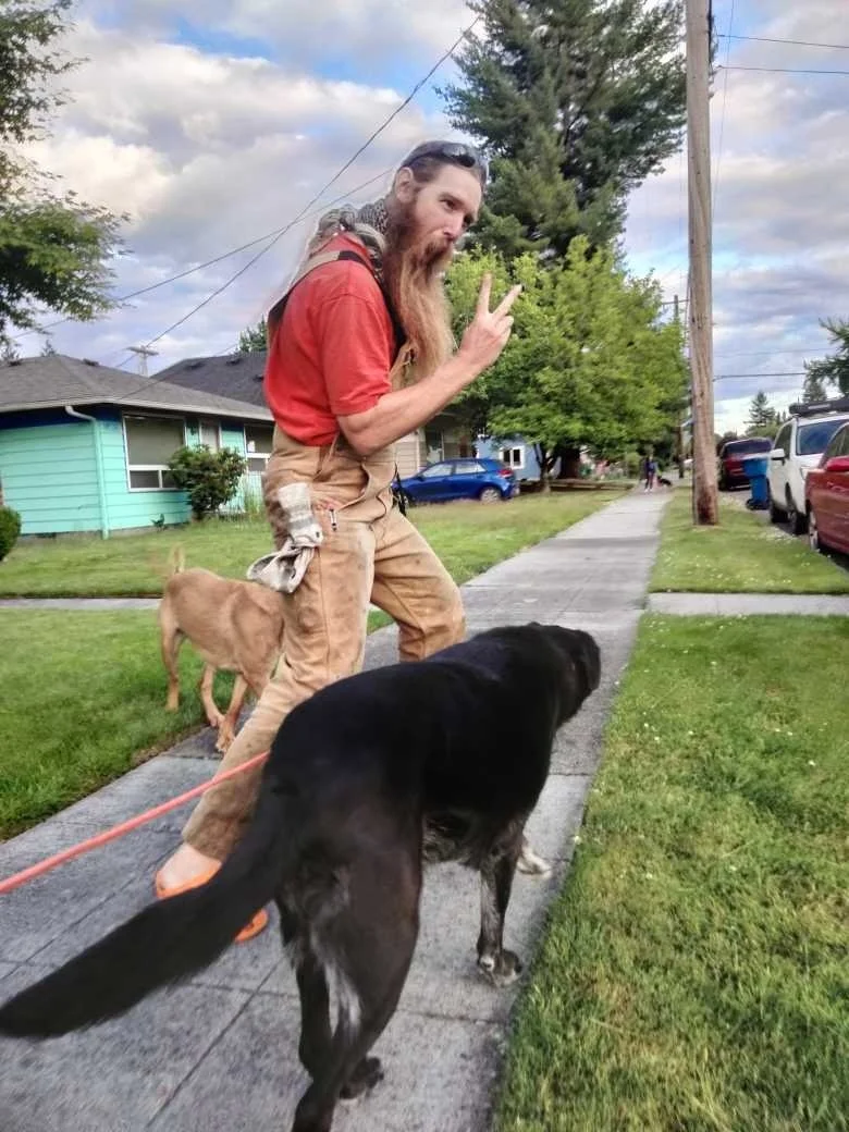 A man with a long beard making a peace sign and posing on a sidewalk with two dogs, one black and one tan, in a suburban neighborhood with houses, trees, and parked cars.