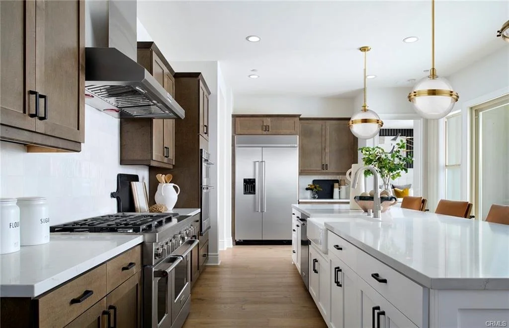 Modern kitchen with brown and white cabinets, stainless steel appliances, a large white island with a sink, pendant lights, and large windows.