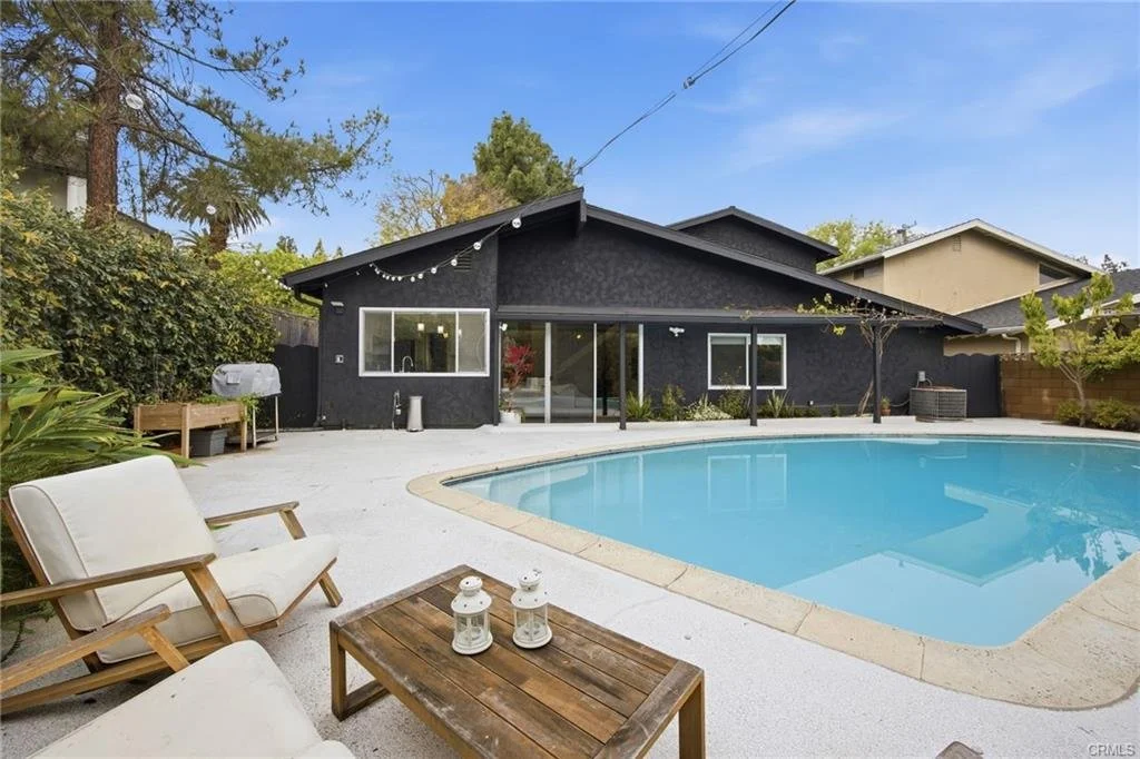 Backyard with a swimming pool, patio furniture including chairs and a small table with lanterns, and a black house with string lights and a patio area.