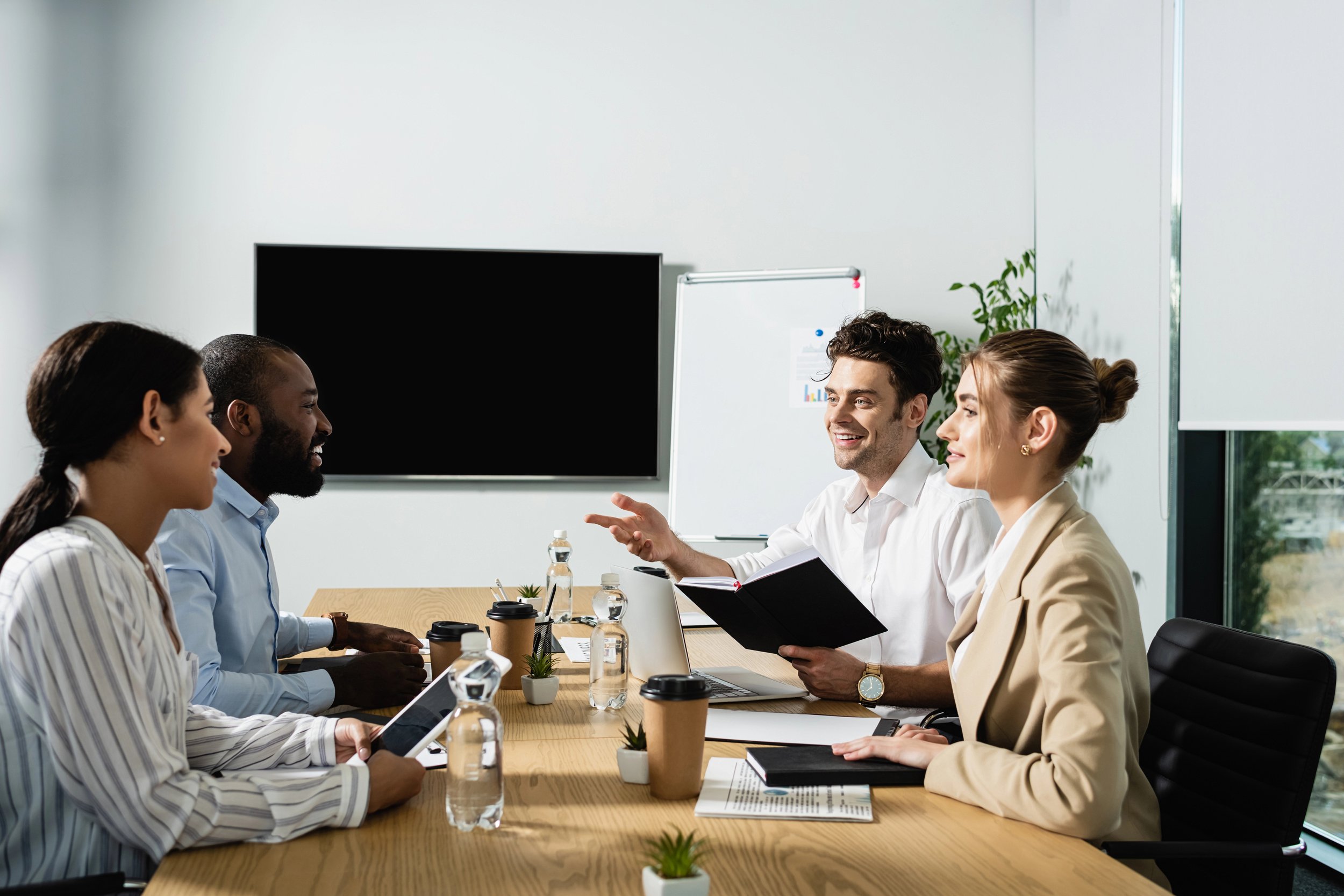 Four people in business attire sitting at a conference table in a modern office, engaged in discussion. One person is speaking, others are listening, with papers, drinks, and a laptop on the table.