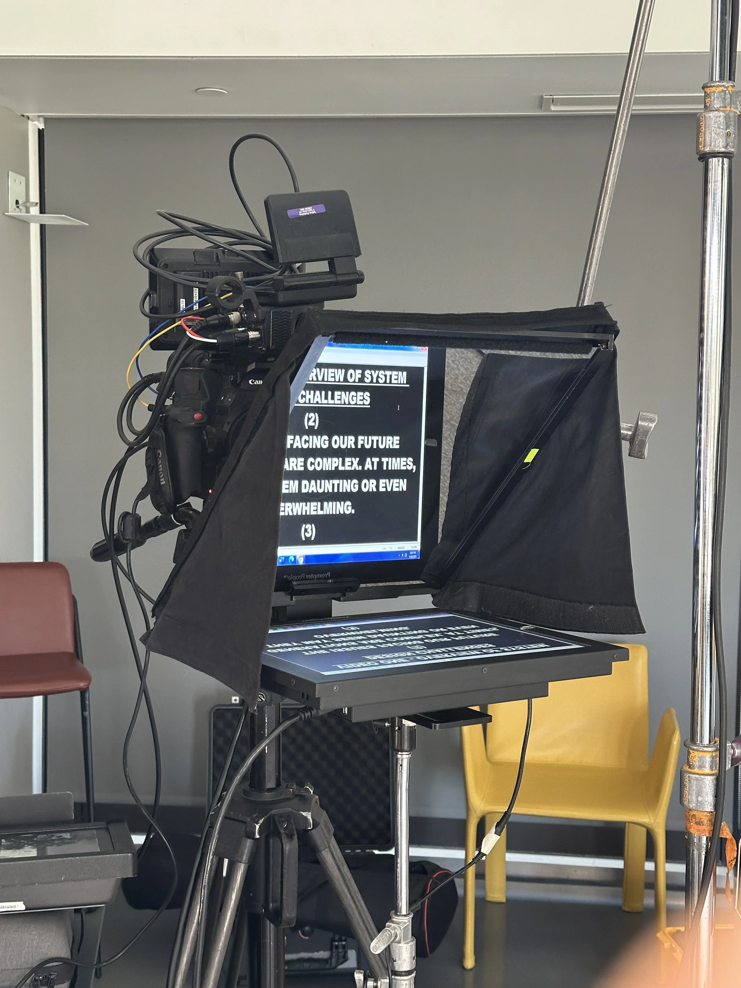 A video camera and monitor setup capturing a presentation slide on a tripod stand in an indoor setting with chairs, including a red chair and a yellow chair, in the background.