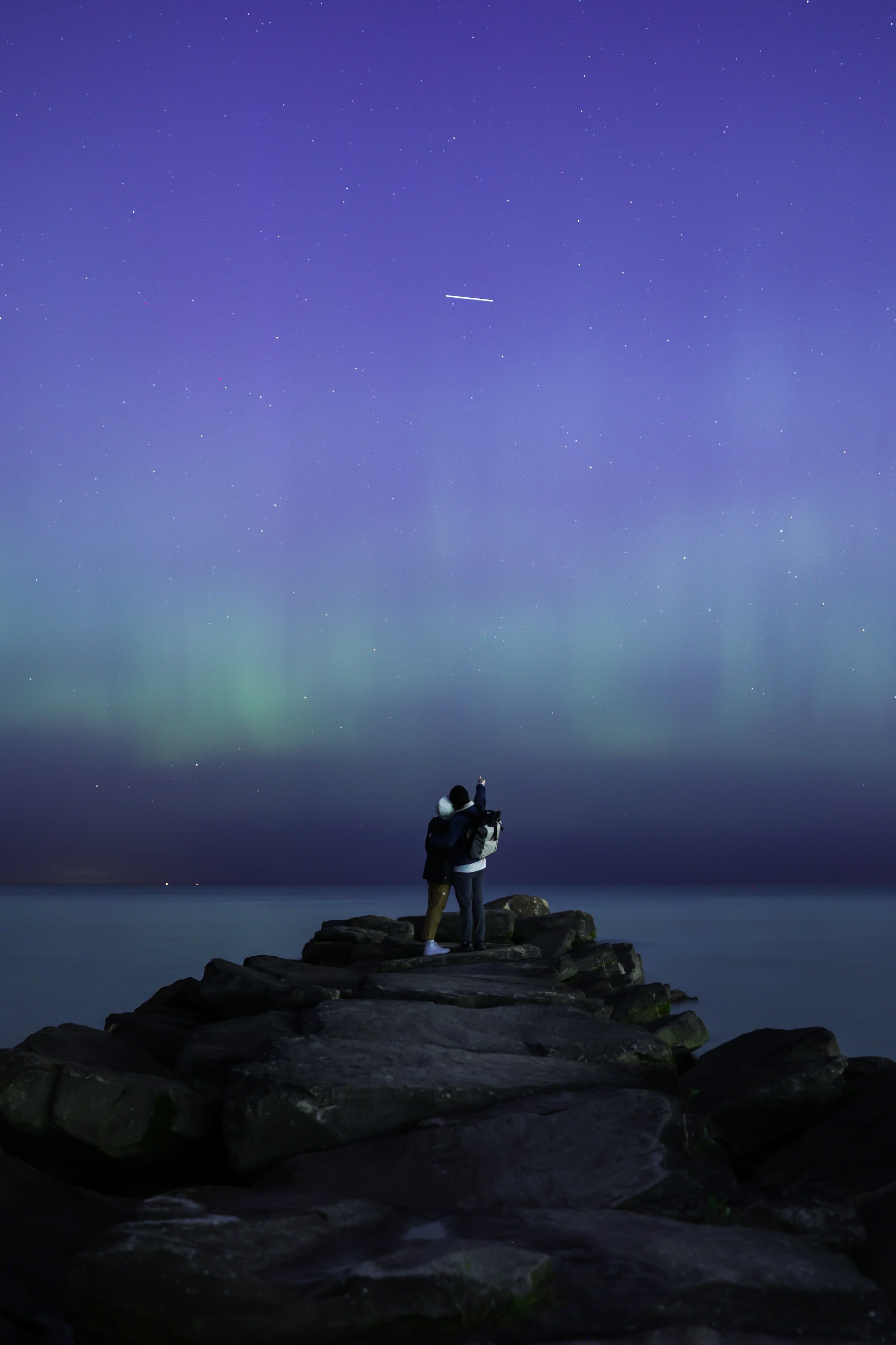 Two people standing on a rocky pier at night, looking up at the star-filled sky and northern lights.