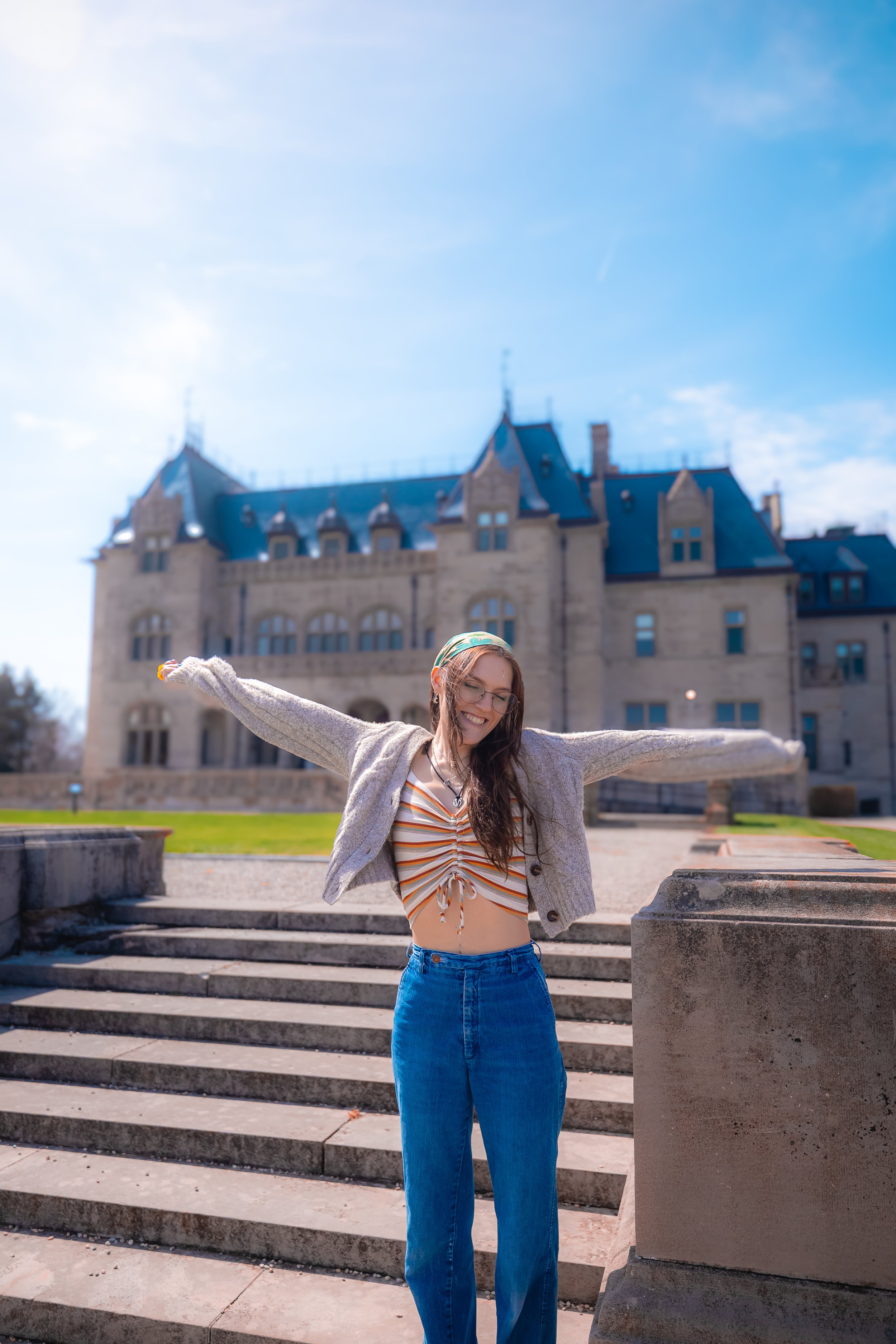 A young woman with long brown hair, glasses, and a headscarf smiling with her arms outstretched in front of a large castle-like building on a sunny day, wearing a striped crop top, a gray sweater, and blue jeans.