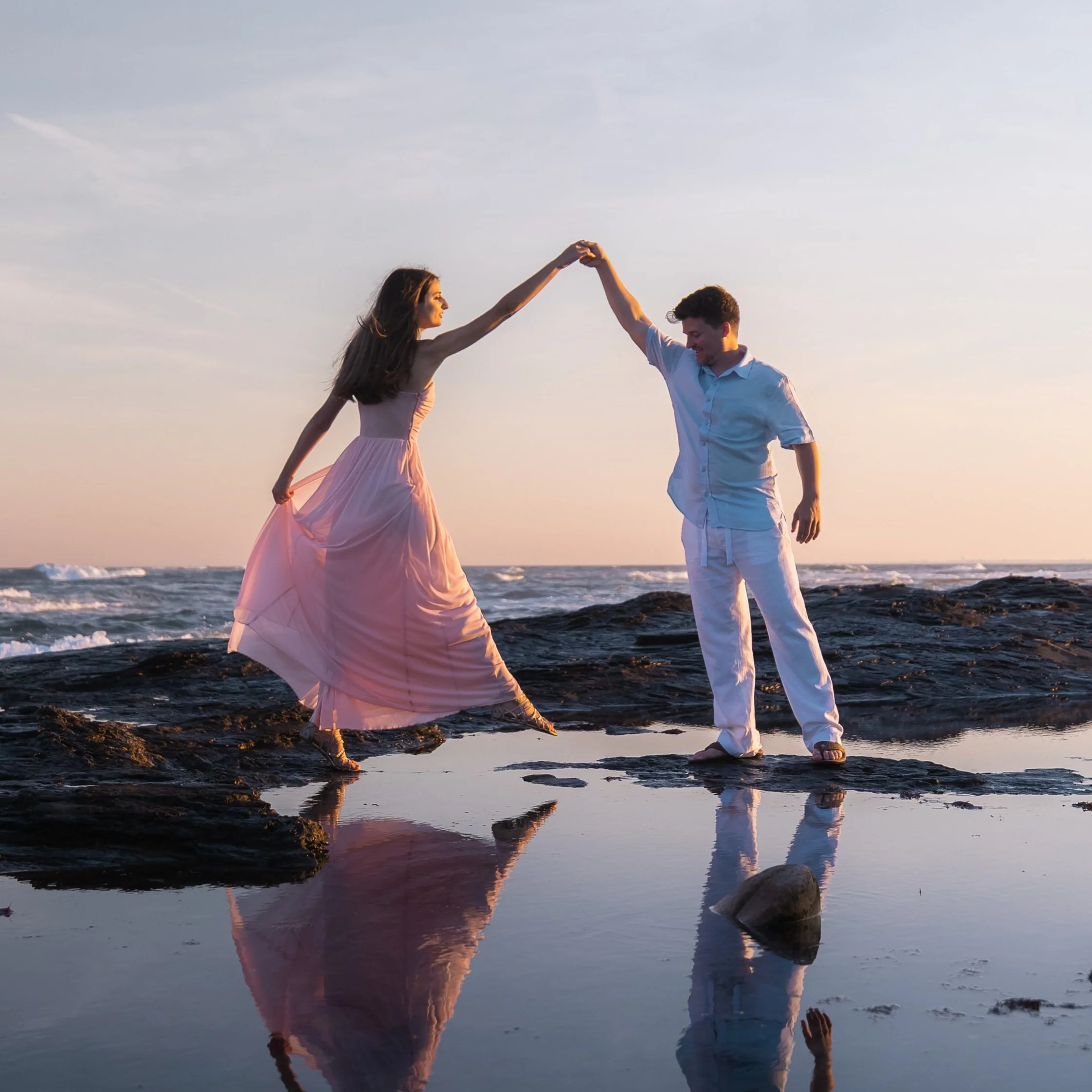 A young couple dancing on rocks by the ocean at sunset, with the woman in a pink dress and the man in white clothing, holding hands and smiling.