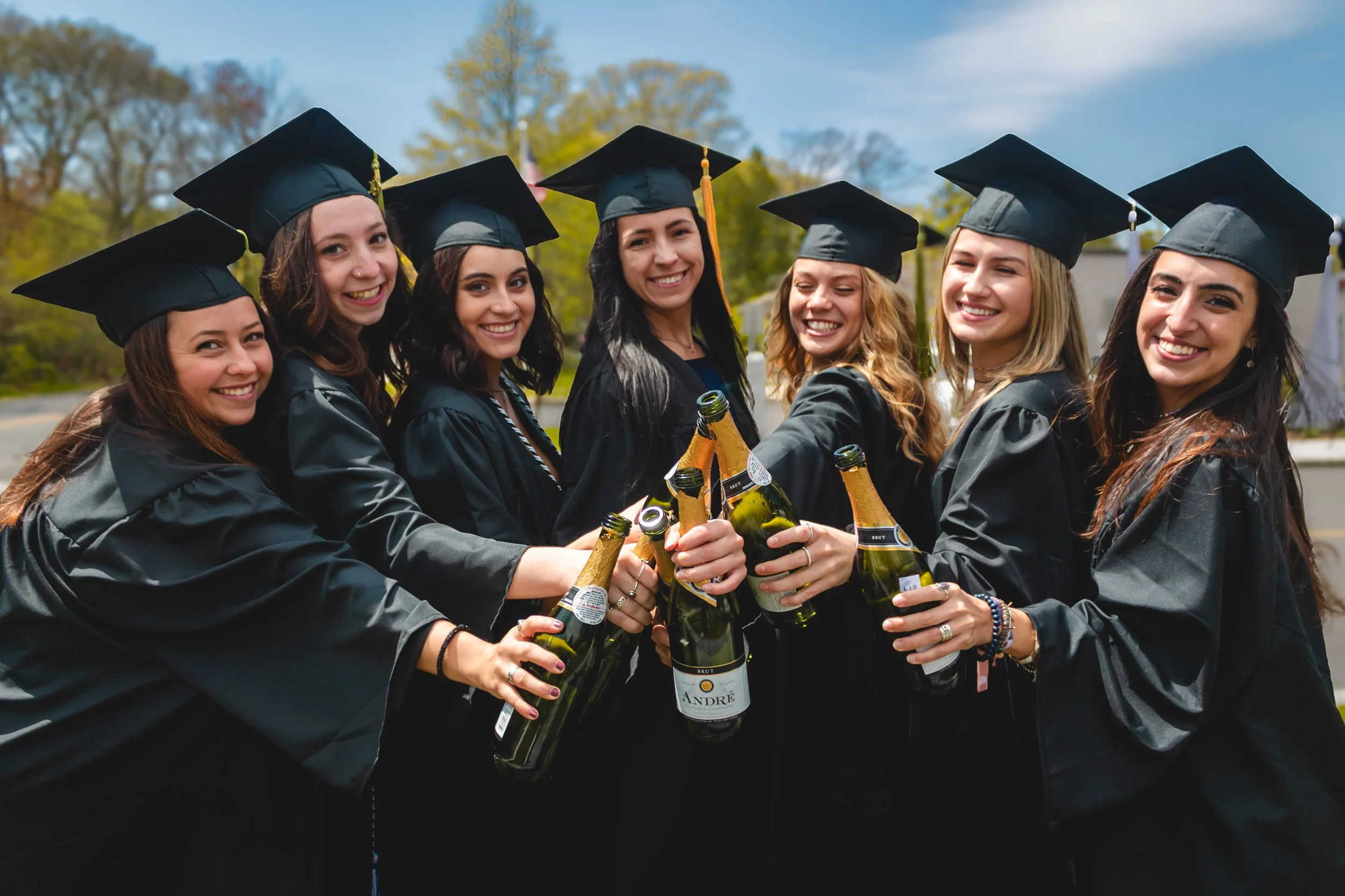 Group of eight young women wearing black graduation caps and gowns, smiling and holding champagne bottles in an outdoor setting.