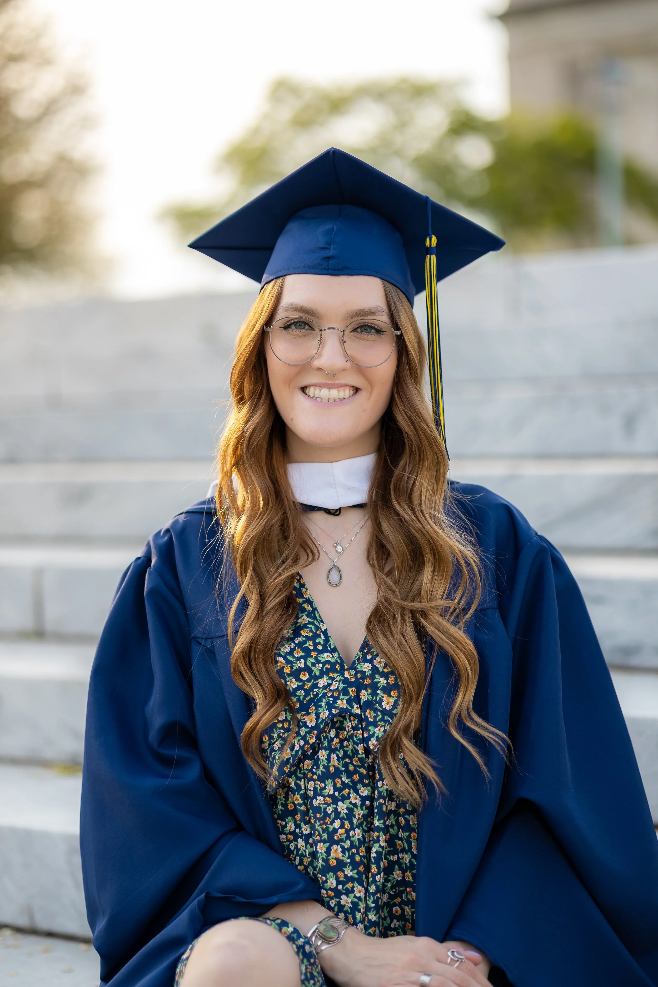 Young woman in a blue graduation cap and gown sitting on steps outside, smiling