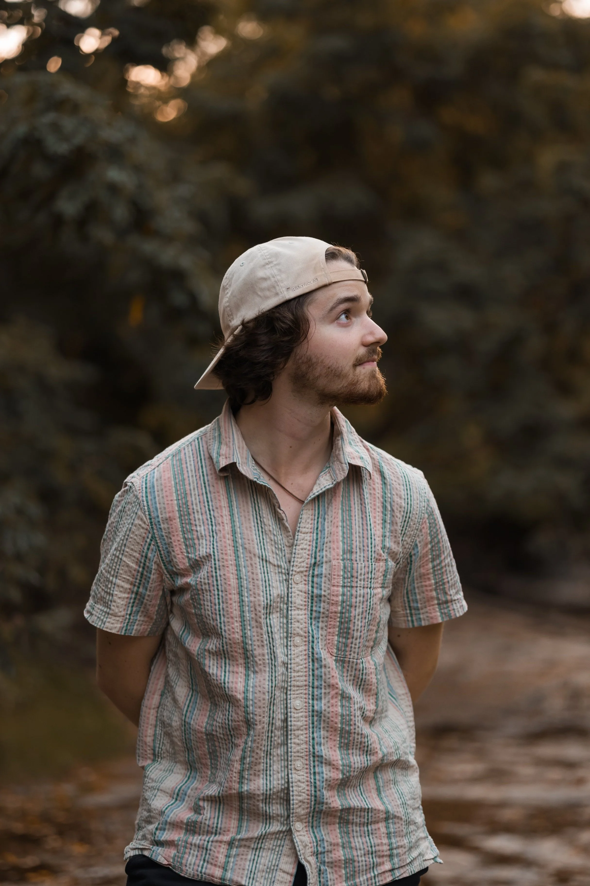 A young man with curly brown hair, a beard, and a beige baseball cap, standing outdoors with trees and a dirt path in the background. He is wearing a short-sleeved, button-up, striped shirt and looking to his right.
