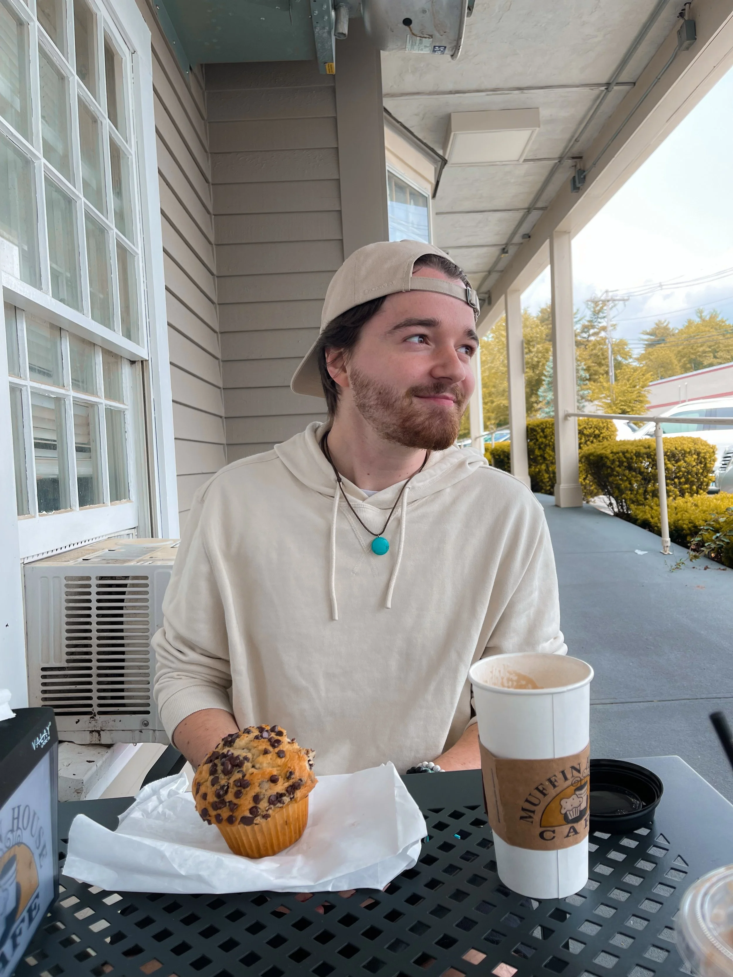 A young man sitting outdoors at a table with a muffin and a cup of coffee, wearing a beige hoodie and a backward baseball cap, looking to his right with a slight smile.